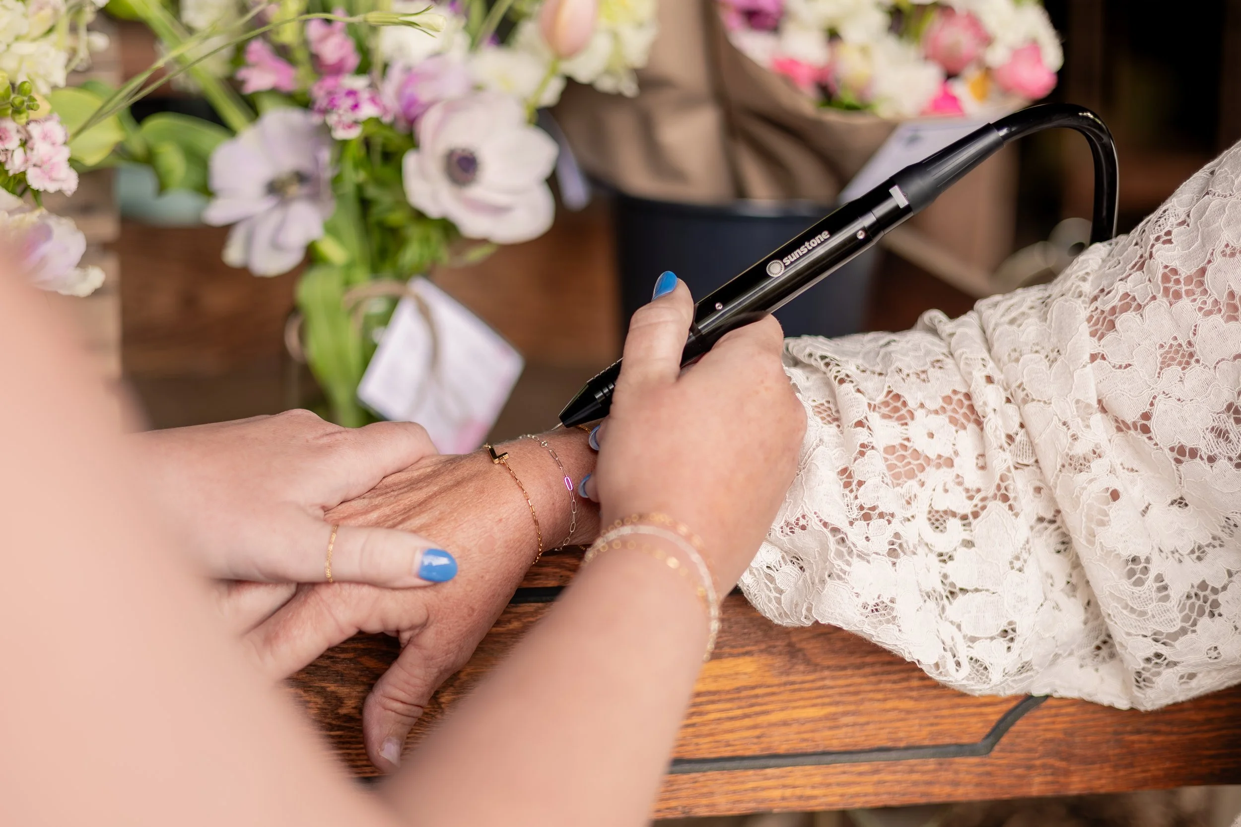 A person with blue painted nails and gold jewelry is holding another person's hand, which has similar jewelry, while sitting at a wooden table with floral arrangements in the background.