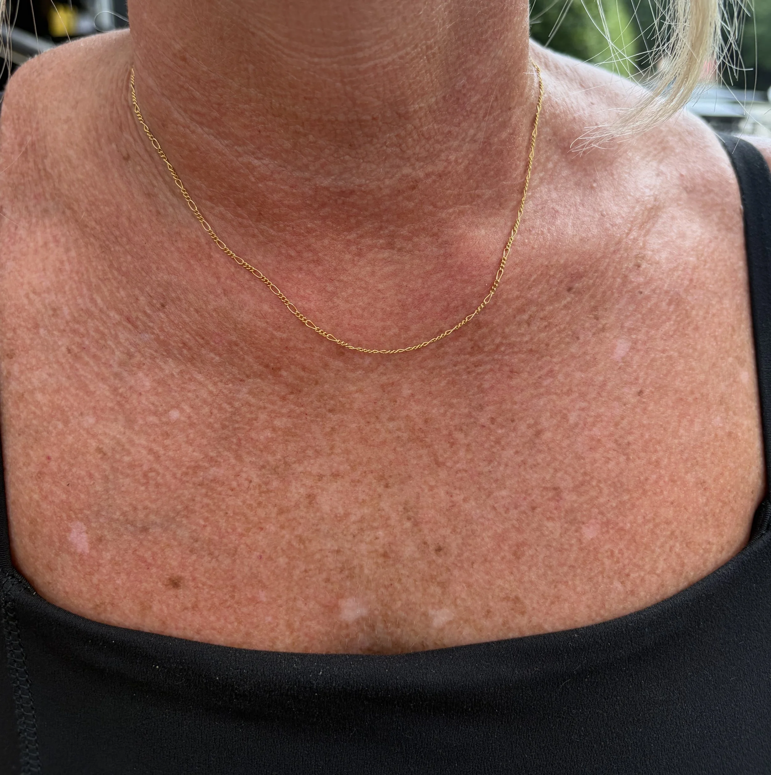 Close-up of a woman's neck and shoulder showing skin with some freckles, wearing a delicate gold chain necklace and a black top.