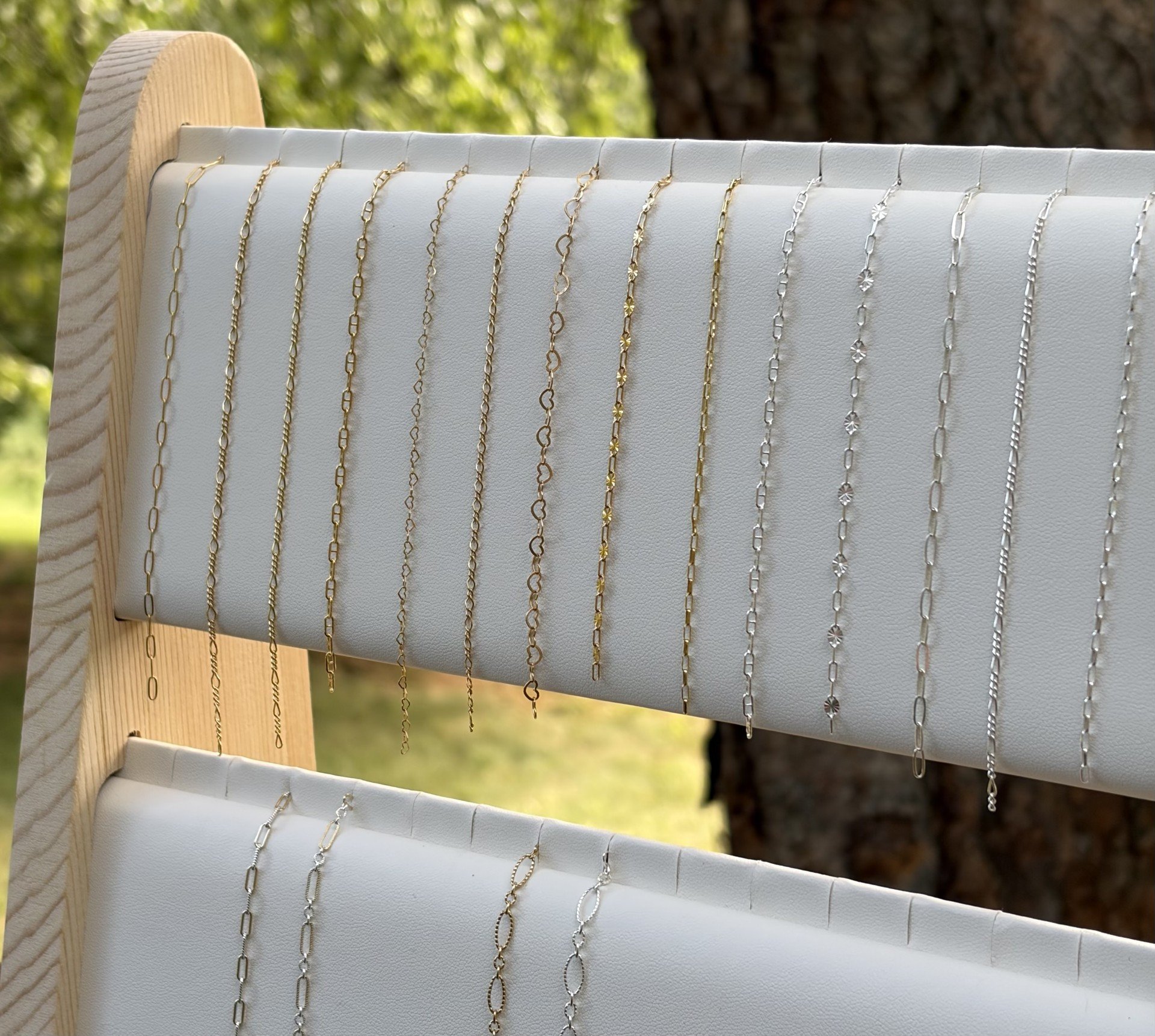 Display of various necklaces in gold and silver, arranged on a white padded jewelry stand outdoors with blurred green foliage and tree trunk in the background.