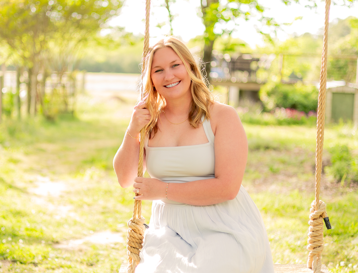 A young woman with blonde hair smiling while sitting on a wooden swing outdoors in a grassy area with trees in the background.