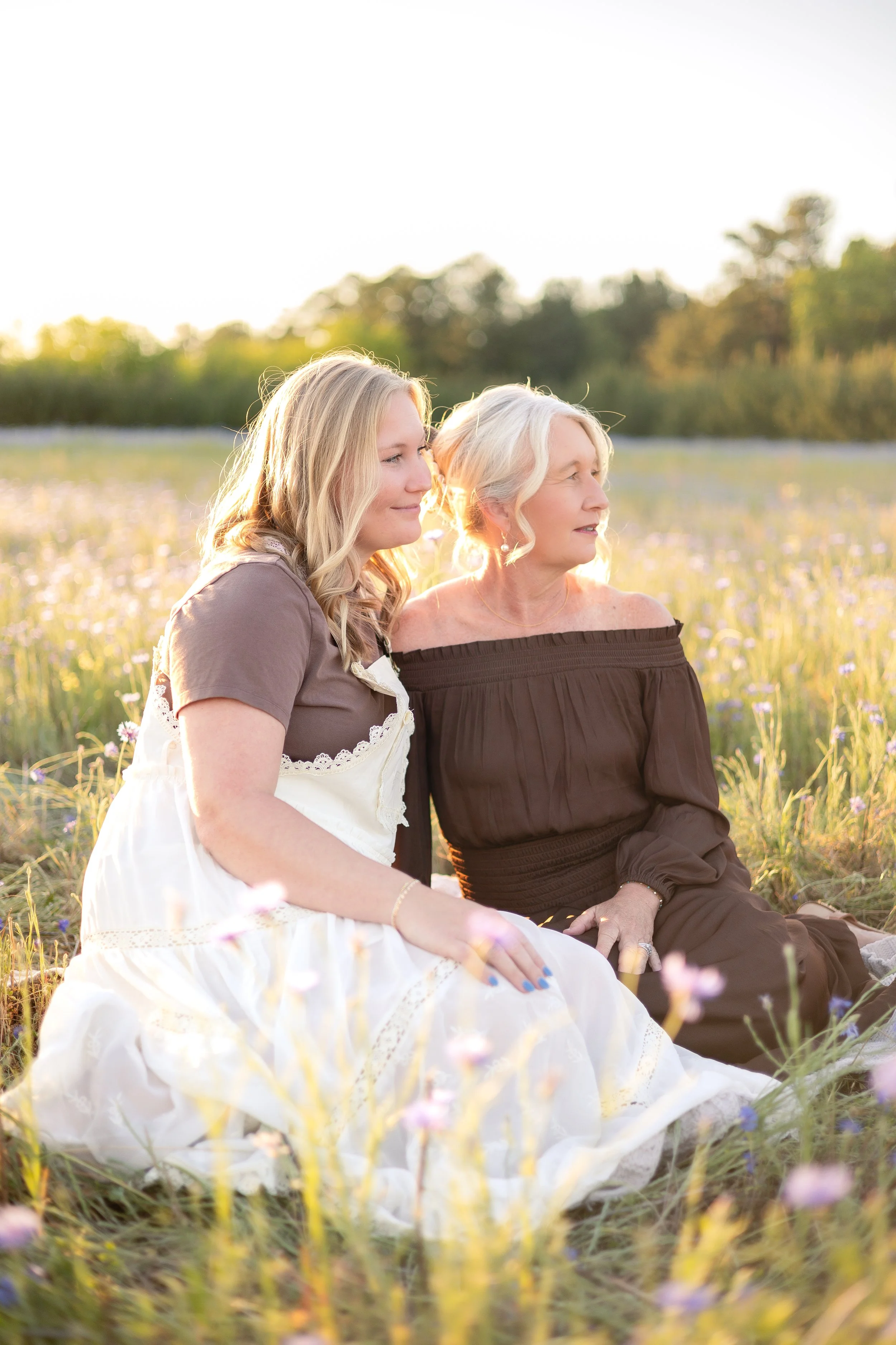 A young woman and an older woman sit together in a field of wildflowers during sunset, enjoying a peaceful moment in nature.
