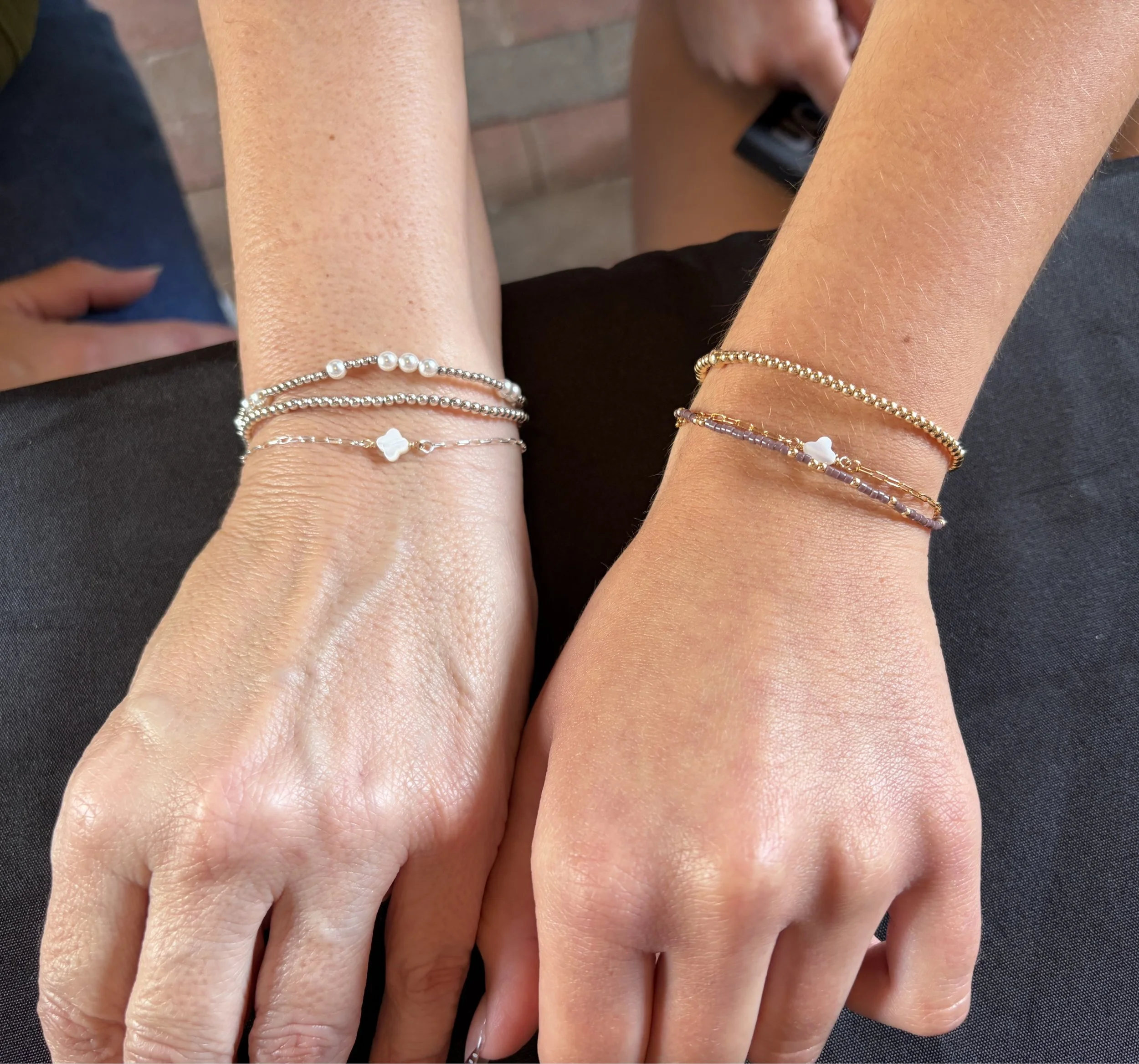 Close-up of two hands with several delicate gold and silver bracelets, one hand wearing a pearl bracelet and a small white charm, with a brick background.