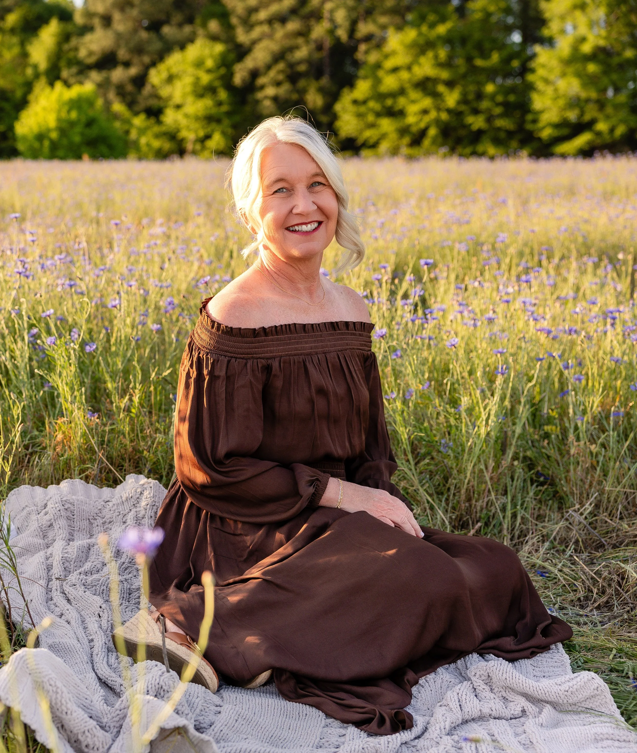 A smiling elderly woman with short gray hair sitting on a white knitted blanket in a field of purple flowers, with green trees in the background, during sunset.