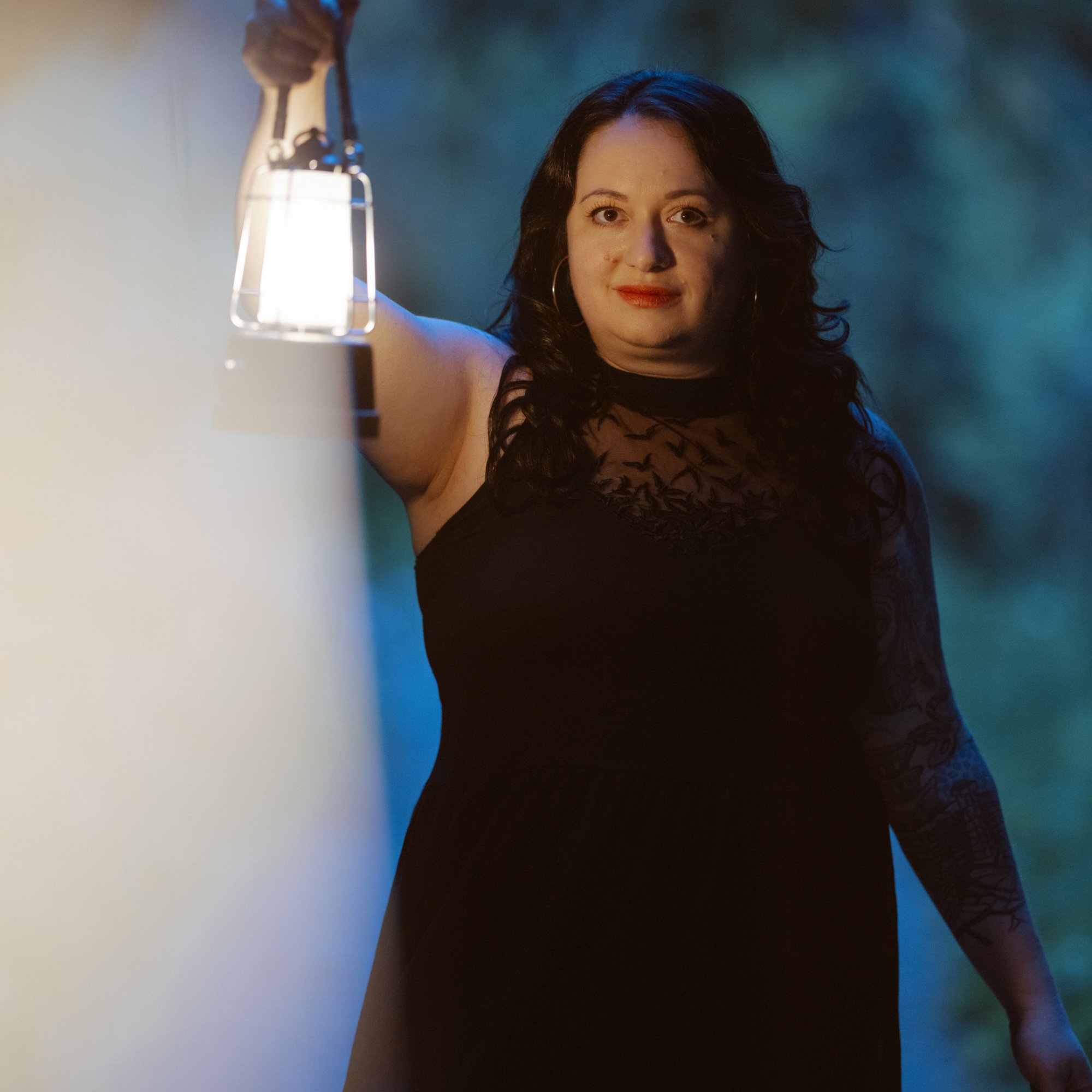 A woman with dark curly hair and earrings smiling in front of a blue and green background, wearing a black lace dress, with a lit lamp in the foreground.