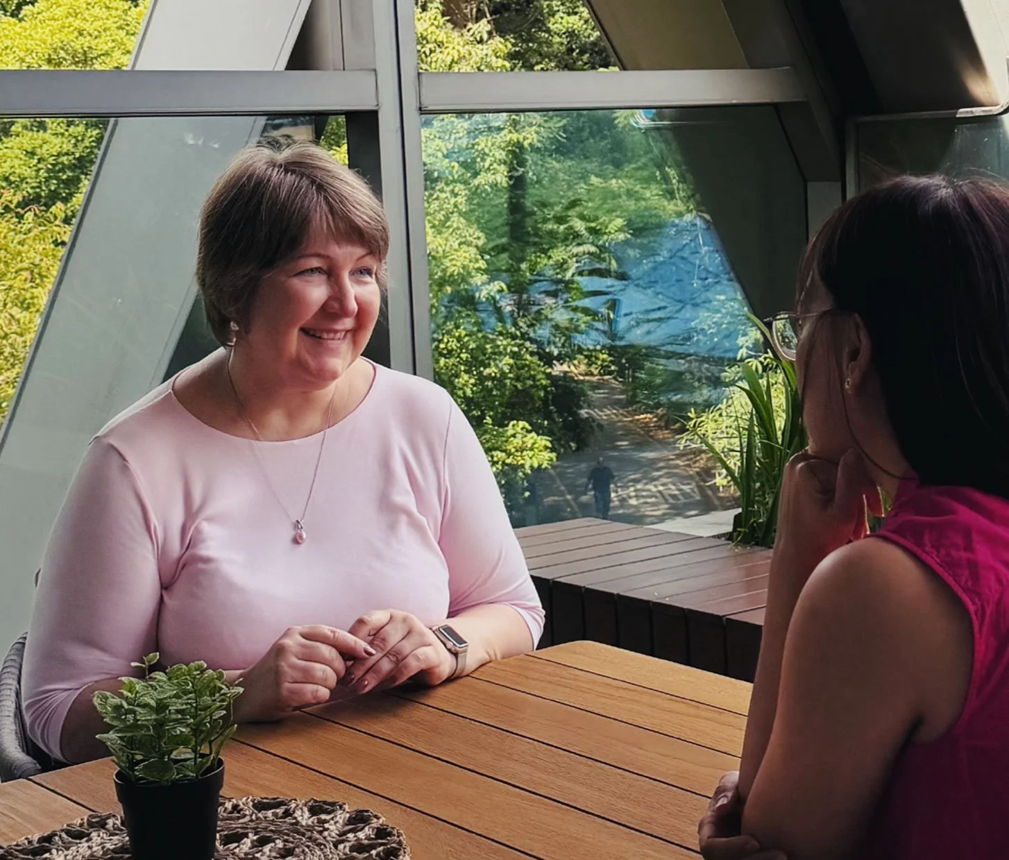 A photo of Sasha facing the camera, at an outdoor table. She is in conversation with a female coachee, who faces away from the camera.