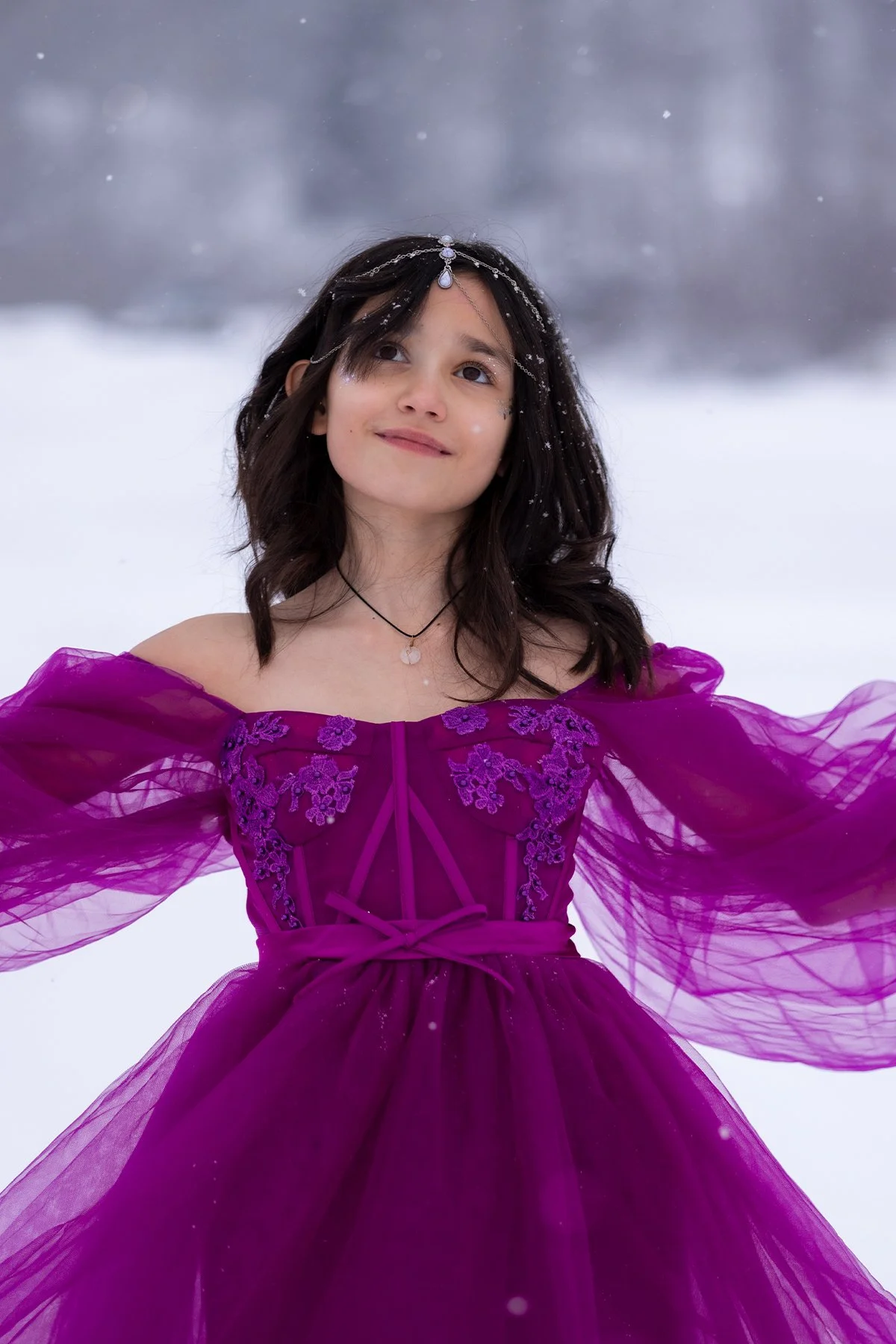 A young woman in a vibrant purple princess dress with sheer, puffed sleeves, standing in a snowy outdoor setting with snow falling around her, looking upward with a gentle smile, wearing a delicate headpiece and jewelry.