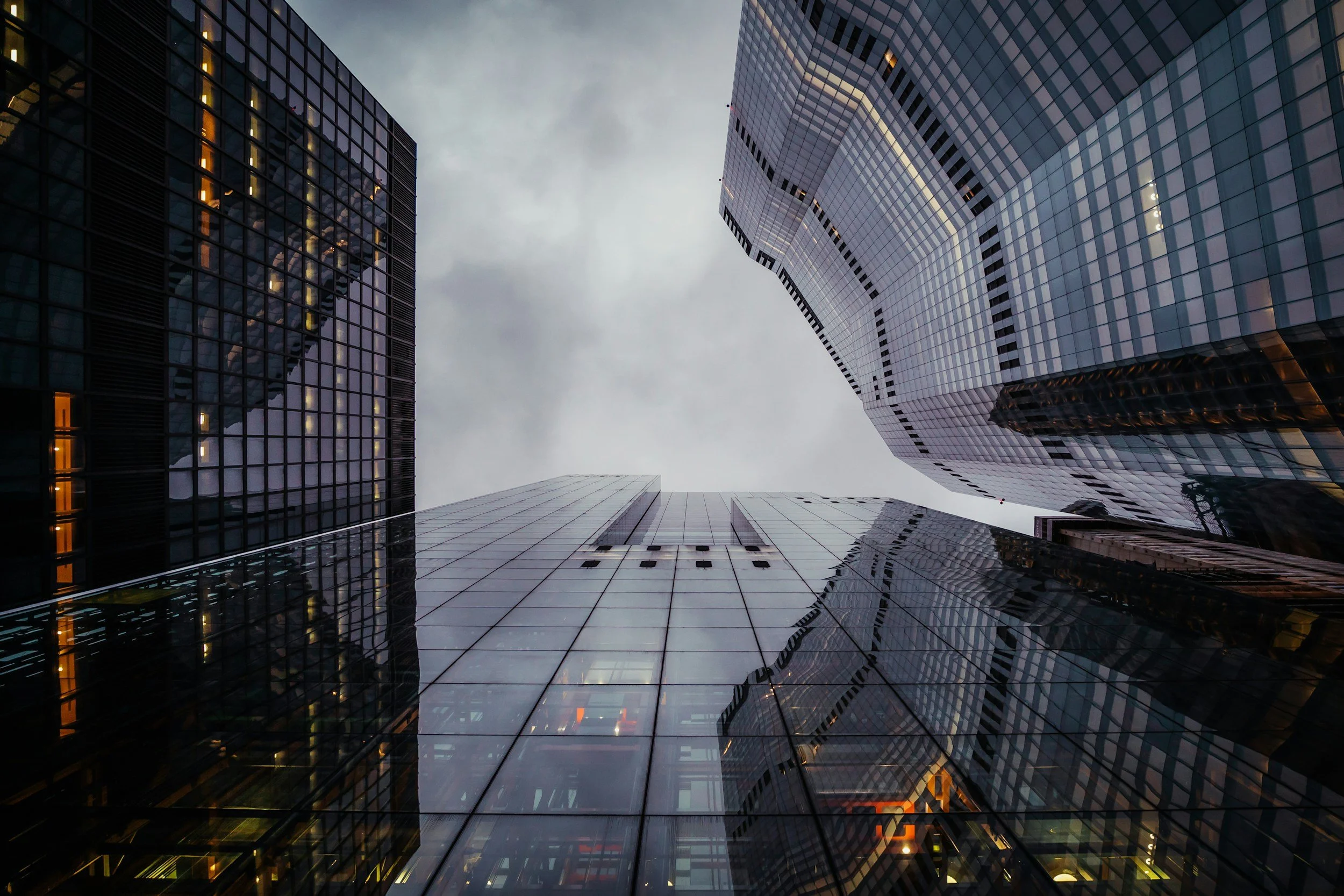A view looking up at tall modern skyscrapers with glass facades reflecting the cloudy sky.