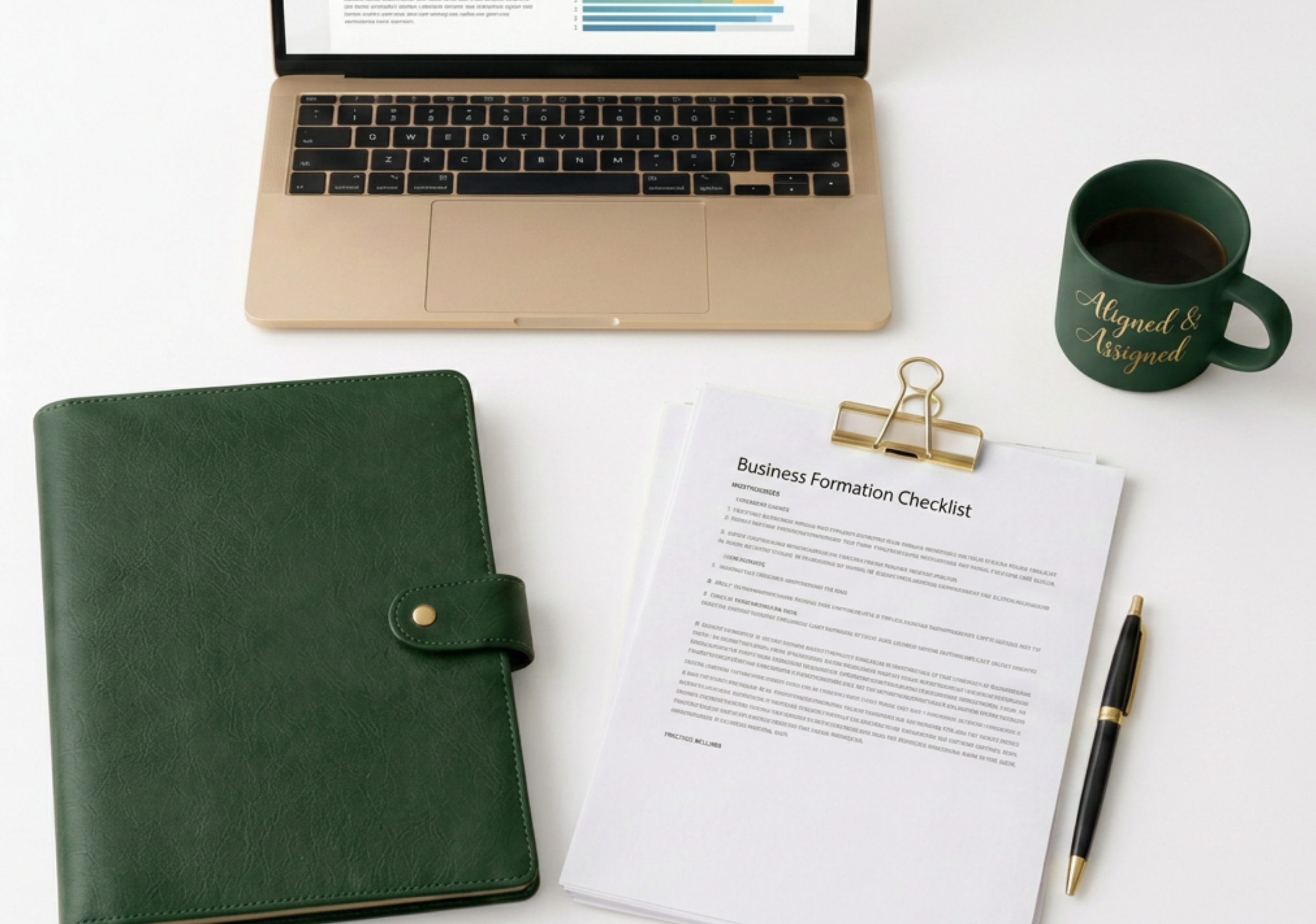 A workspace featuring a laptop with a graph displayed on the screen, a green notebook, a document titled 'Business Formation Checklist', a black pen with gold accents, a gold paperclip, a green coffee mug with the phrase 'Aligned & Assigned' written in gold, and some white papers on a white surface.