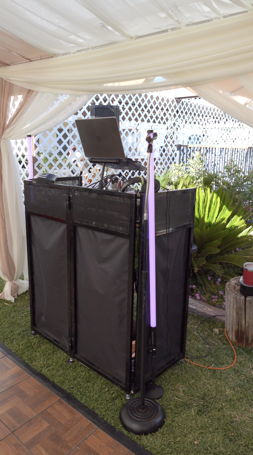 DJ setup with laptop, speakers, microphone, and lighting in a decorated outdoor tent with plants and patterned fencing.