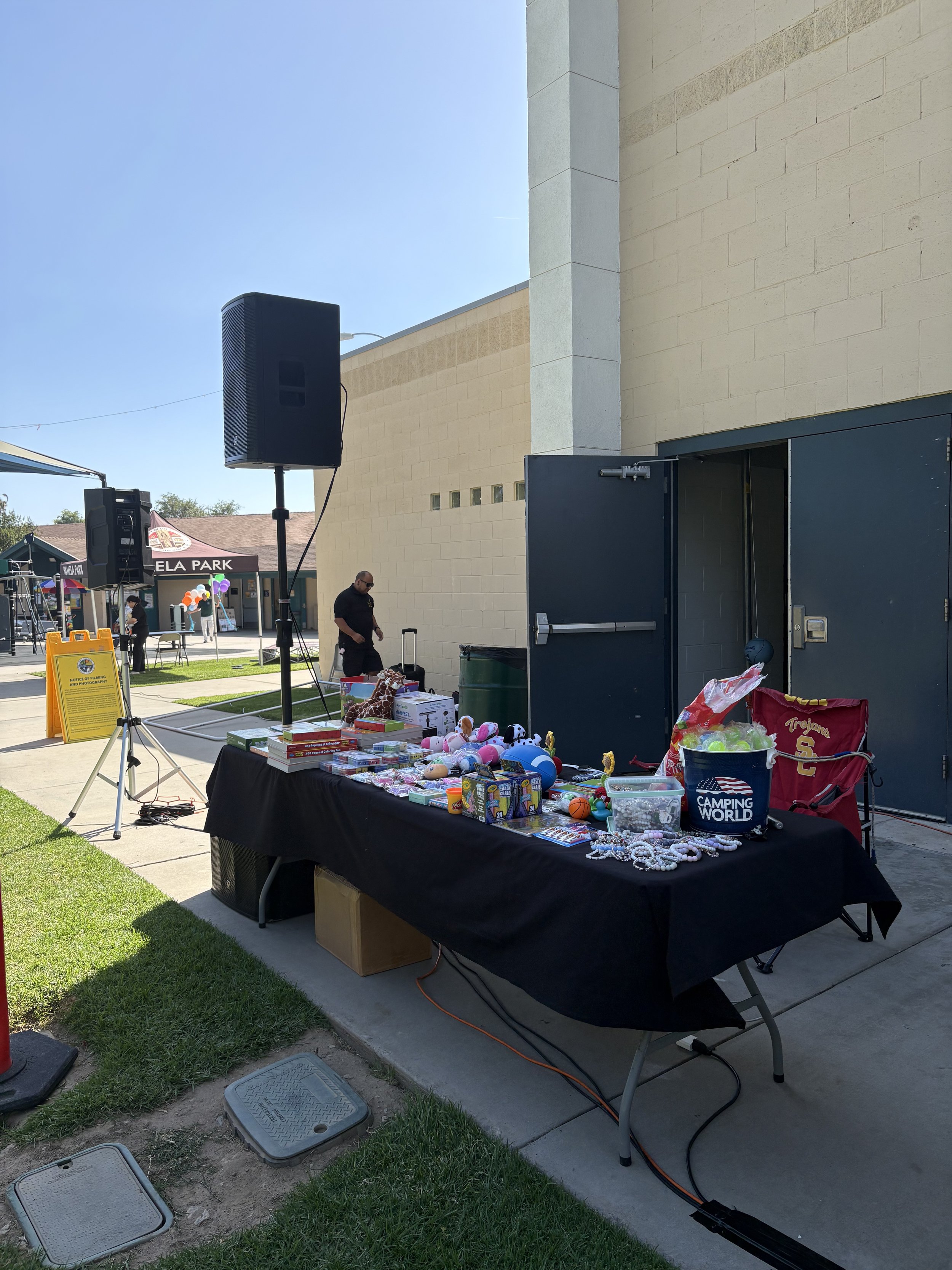 An outdoor yard sale stall with a black tablecloth displaying toys, plush animals, and various items. A man wearing black clothing is near the table. In the background, there are tents, a sign, and a building wall.