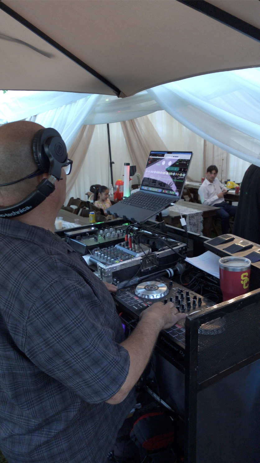 A DJ wearing headphones operates DJ equipment at an event under a tent with white drapes, and people are sitting at tables in the background.