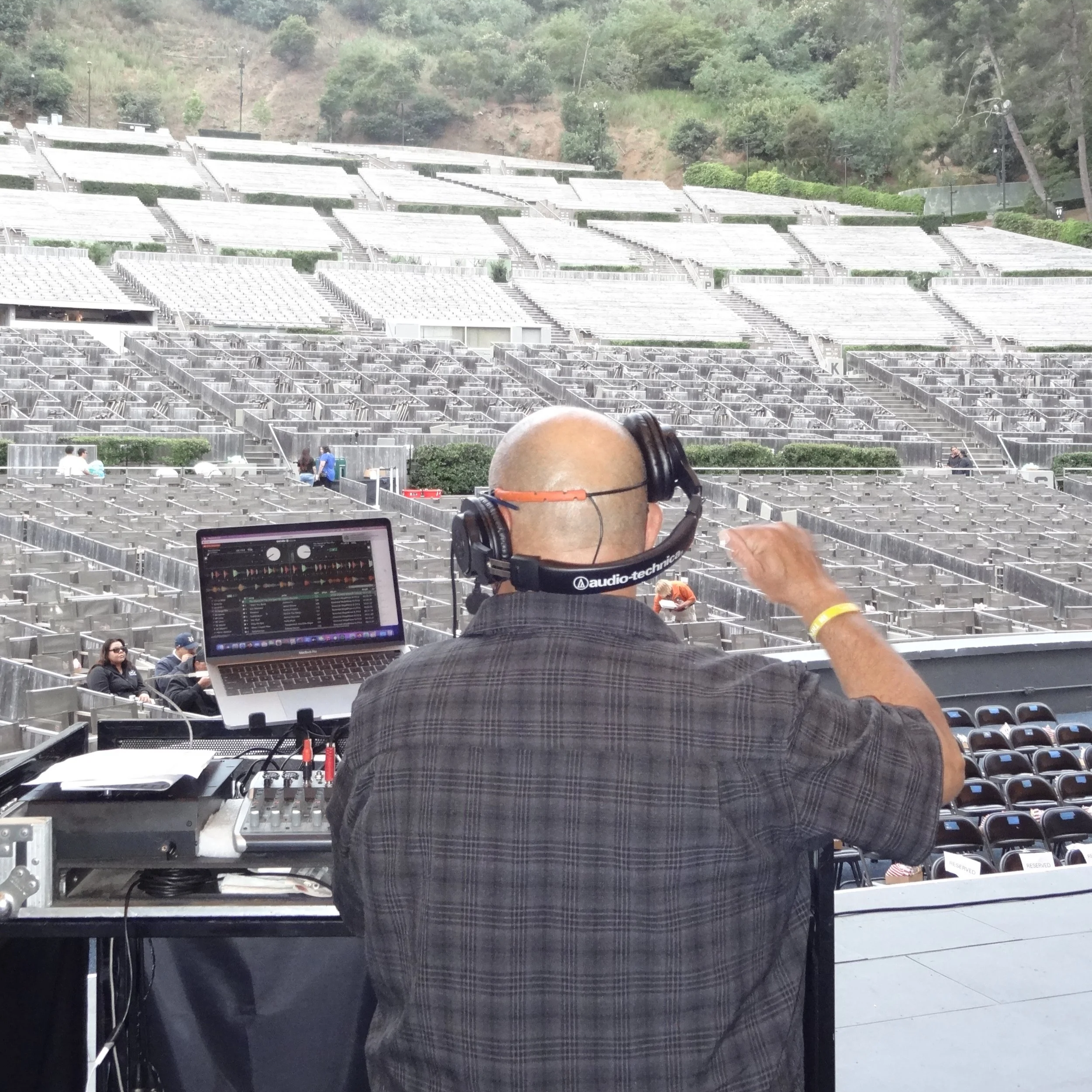 DJ or sound engineer at a music event wearing headphones in front of a DJ controller, in a large outdoor venue with empty tiered seating, trees, and hills in the background.