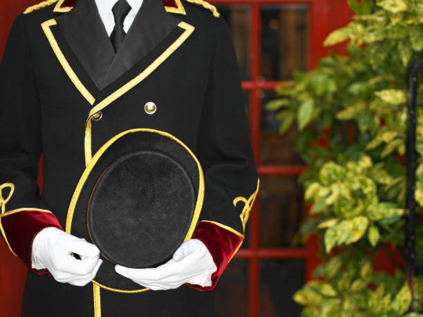 A person in a formal uniform holding a black hat, with a garden and red window in the background.