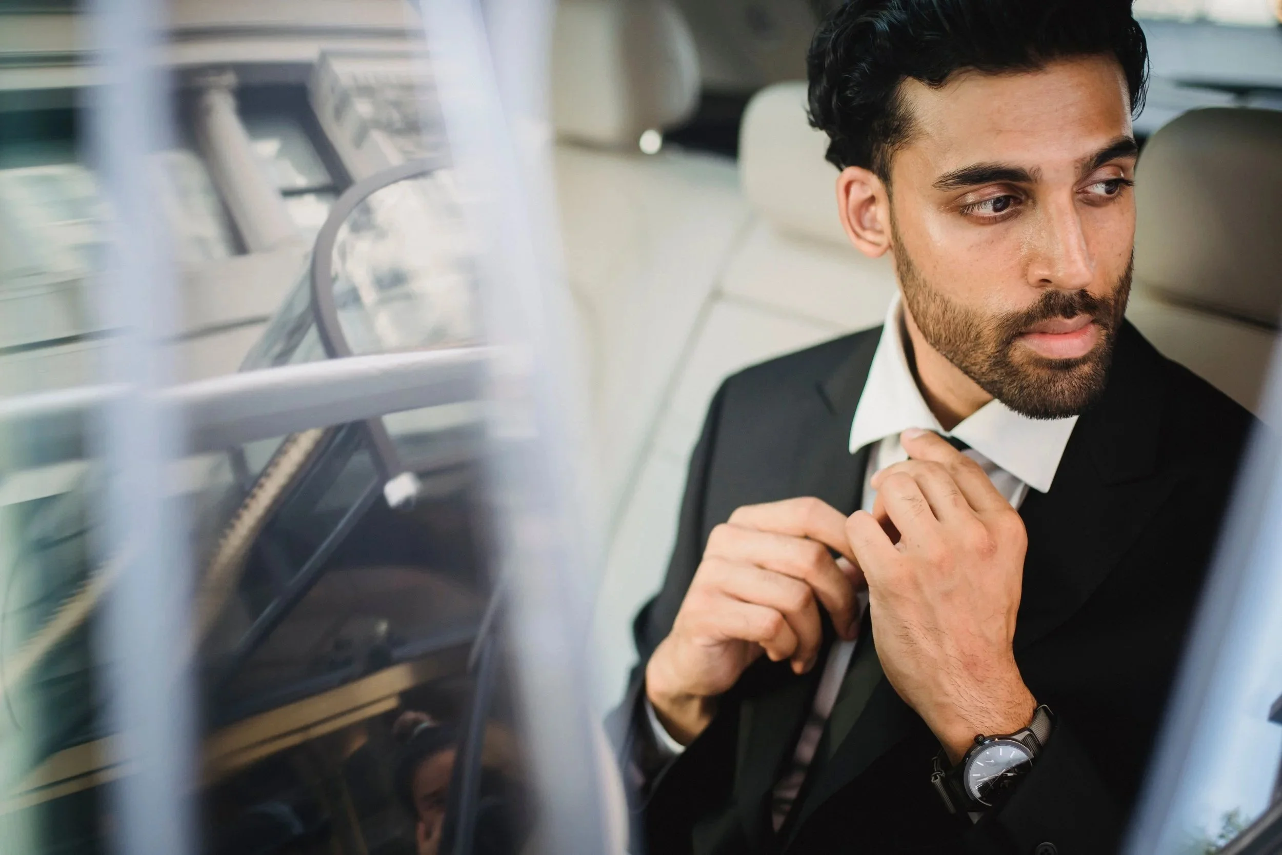 A man in a black suit and bow tie adjusting his collar inside a vehicle, seen through a window with a grid pattern.