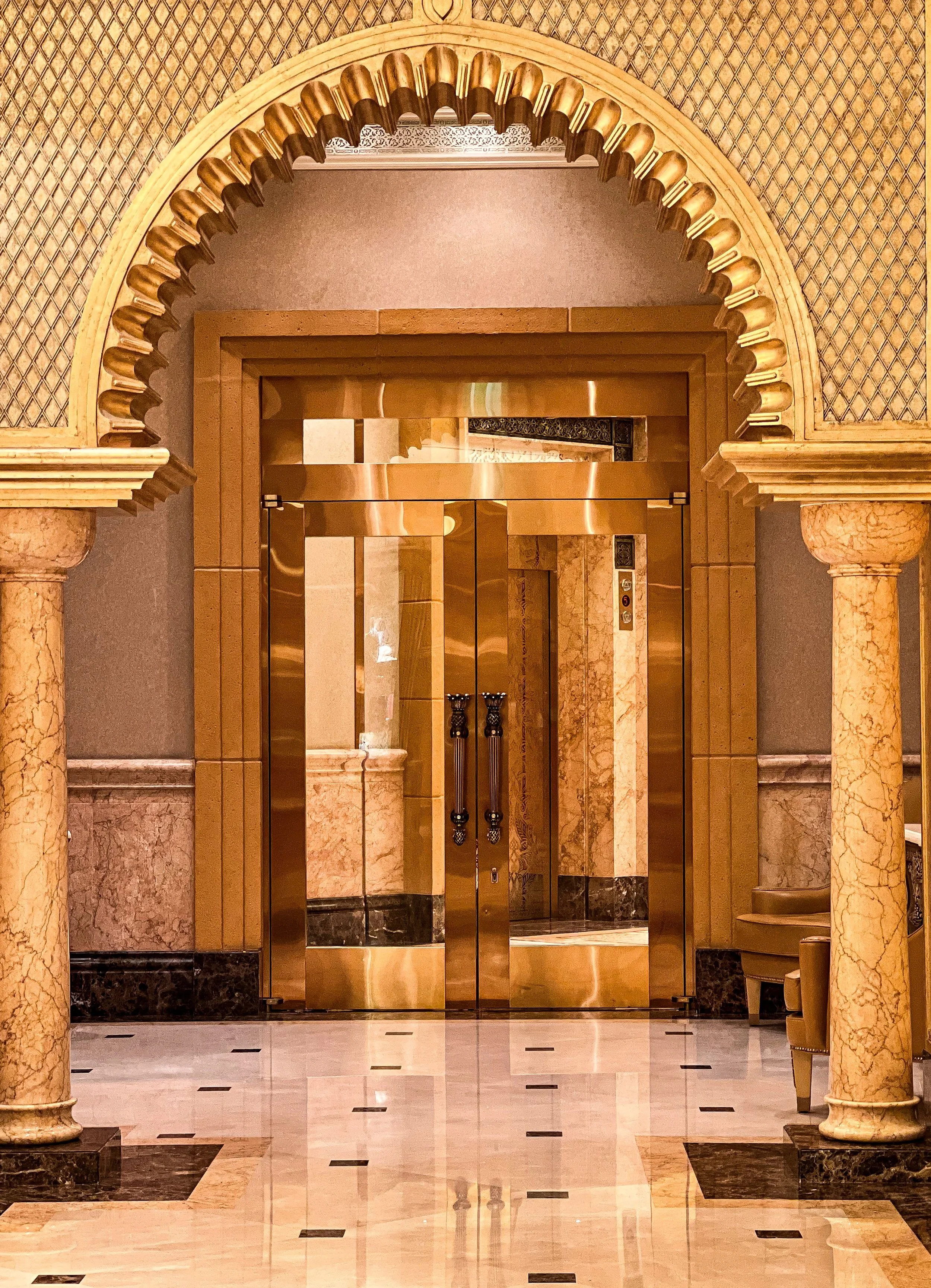 Gold-colored elevator doors surrounded by marble columns and gold decorative arch in a luxurious building lobby.