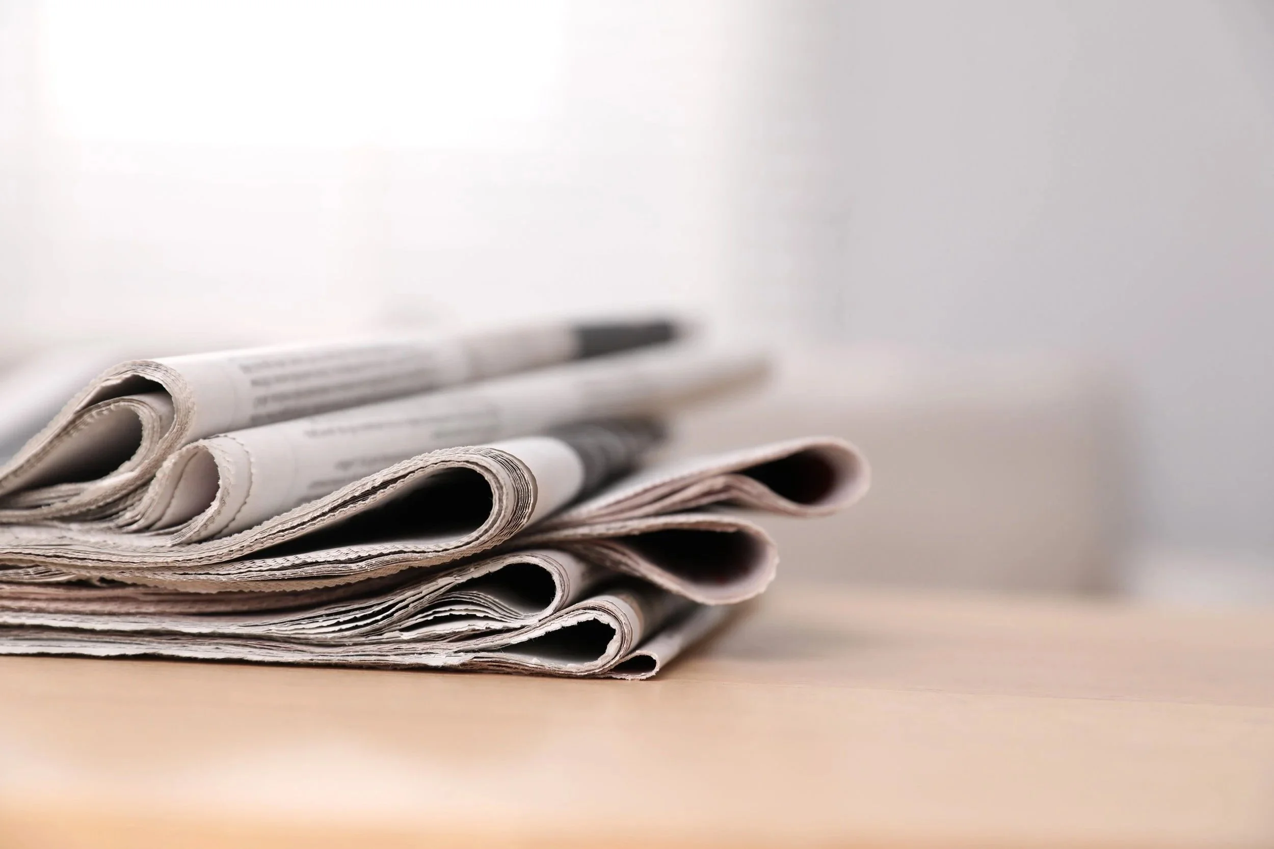 Stack of folded newspapers on a light-colored surface with a plain light-colored wall background.