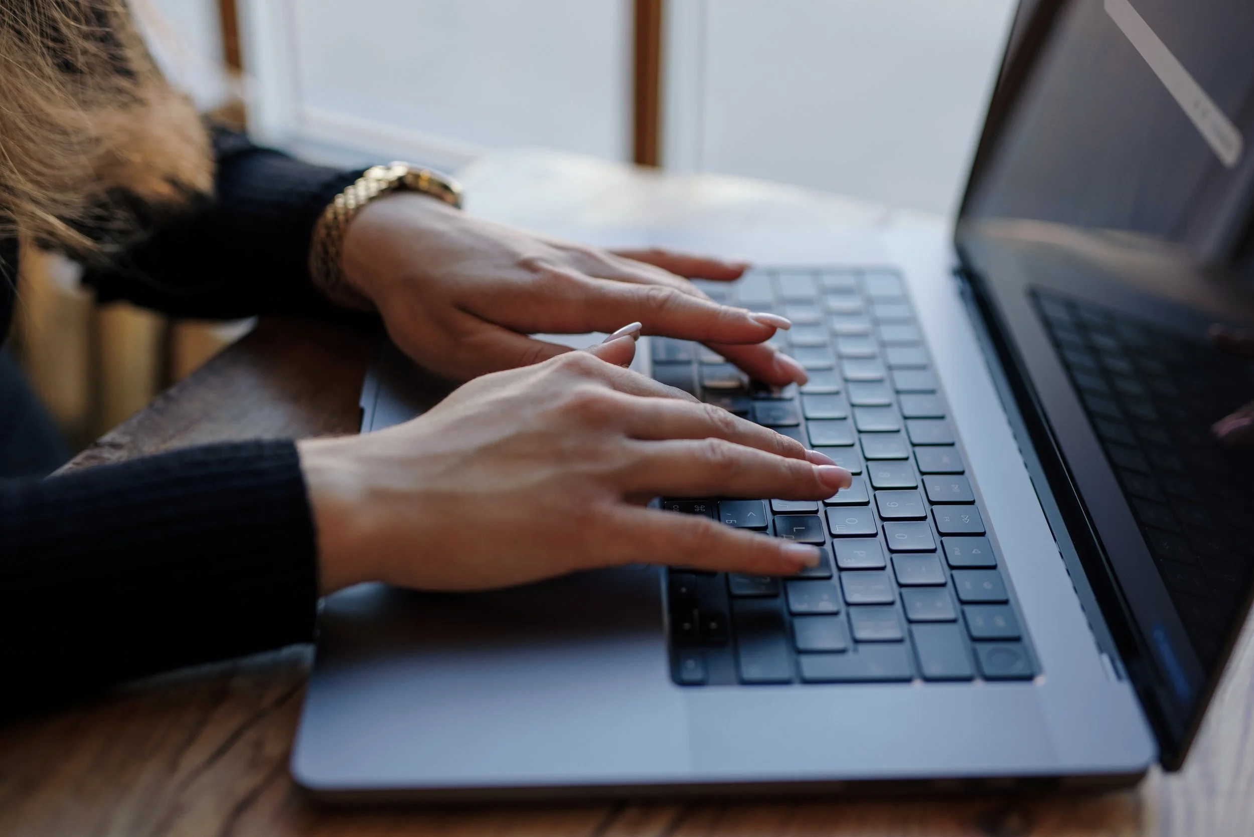 Close-up of a person's hands typing on a laptop keyboard on a wooden table.