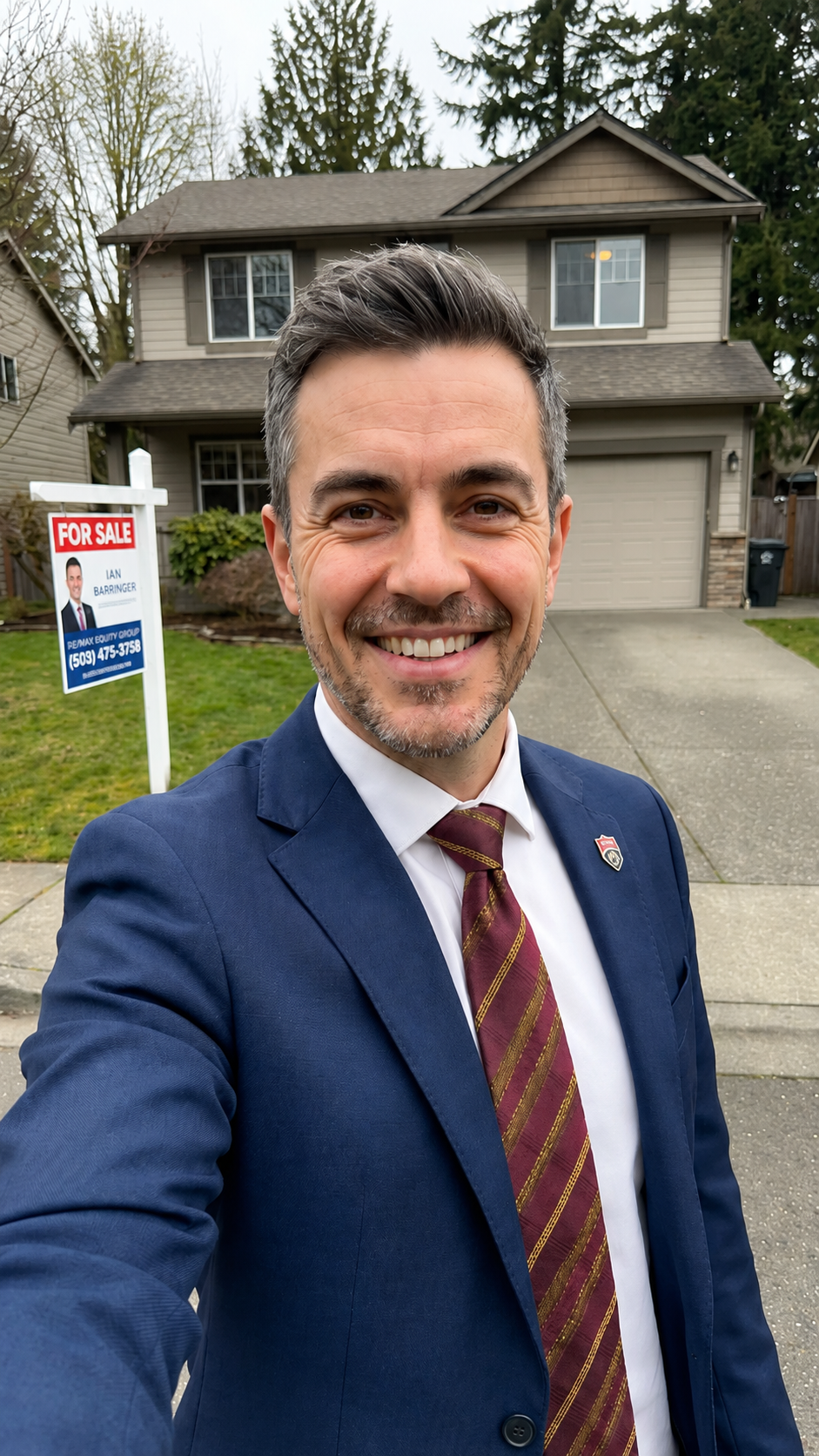 A man in a blue suit and maroon striped tie taking a selfie in front of a two-story house with a "For Sale" sign on the front yard.
