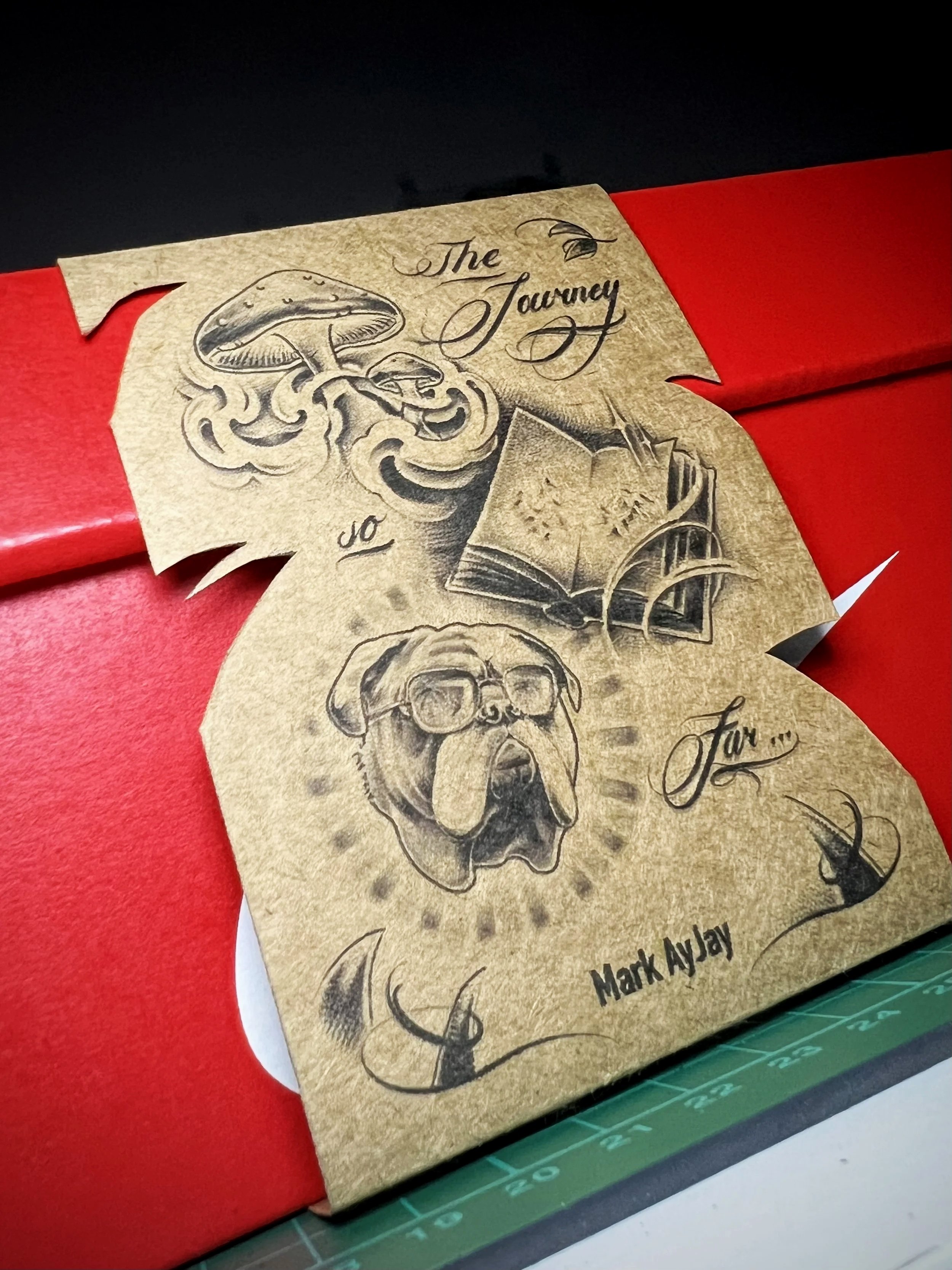 A piece of parchment paper with hand-drawn illustrations of a mushroom, a dog's skull, and a bird, with handwritten text reading "The Journey For..." in cursive, placed on a red surface and a green cutting mat.