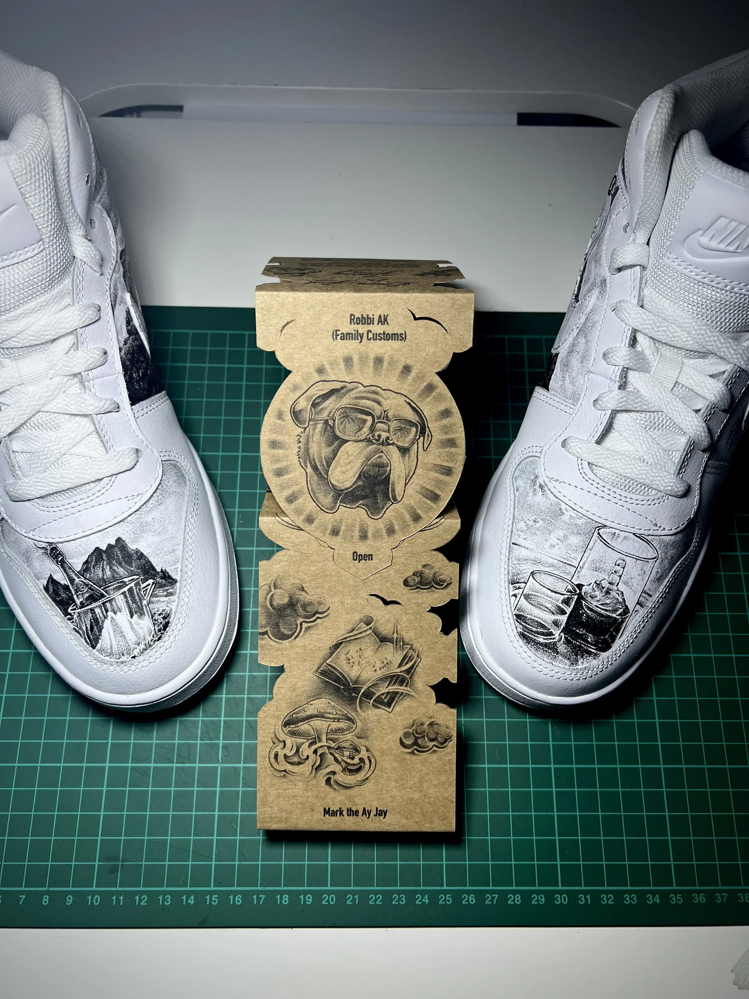 A pair of white Nike sneakers with black and gray design elements, placed on a green cutting mat, with a brown paper tag in between them featuring a dog illustration and the text 'Robbi AK (Family Customs)', 'Open', and 'Mark the Ay Jay'.