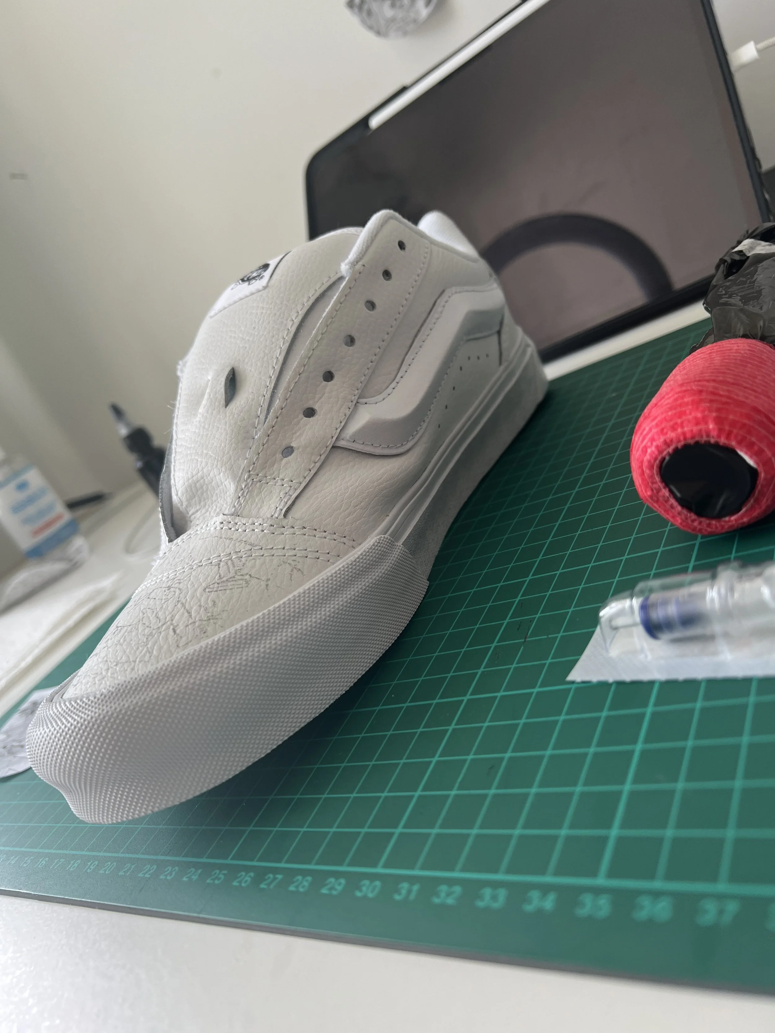 A white sneaker with visible stitching and a textured surface placed on a green cutting mat with a ruler. In the background, there is a computer monitor, a red roll of thread, a black plastic bag, and a plastic package with a syringe on it.
