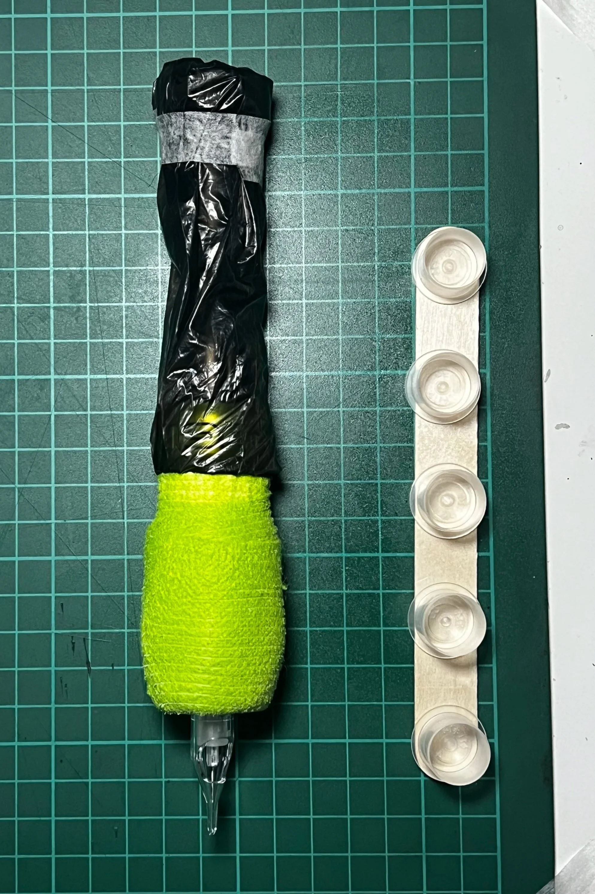A black and green toy gun wrapped in plastic, a strip of tape, a green textured cloth on the handle, with a row of five small transparent plastic cups beside it.