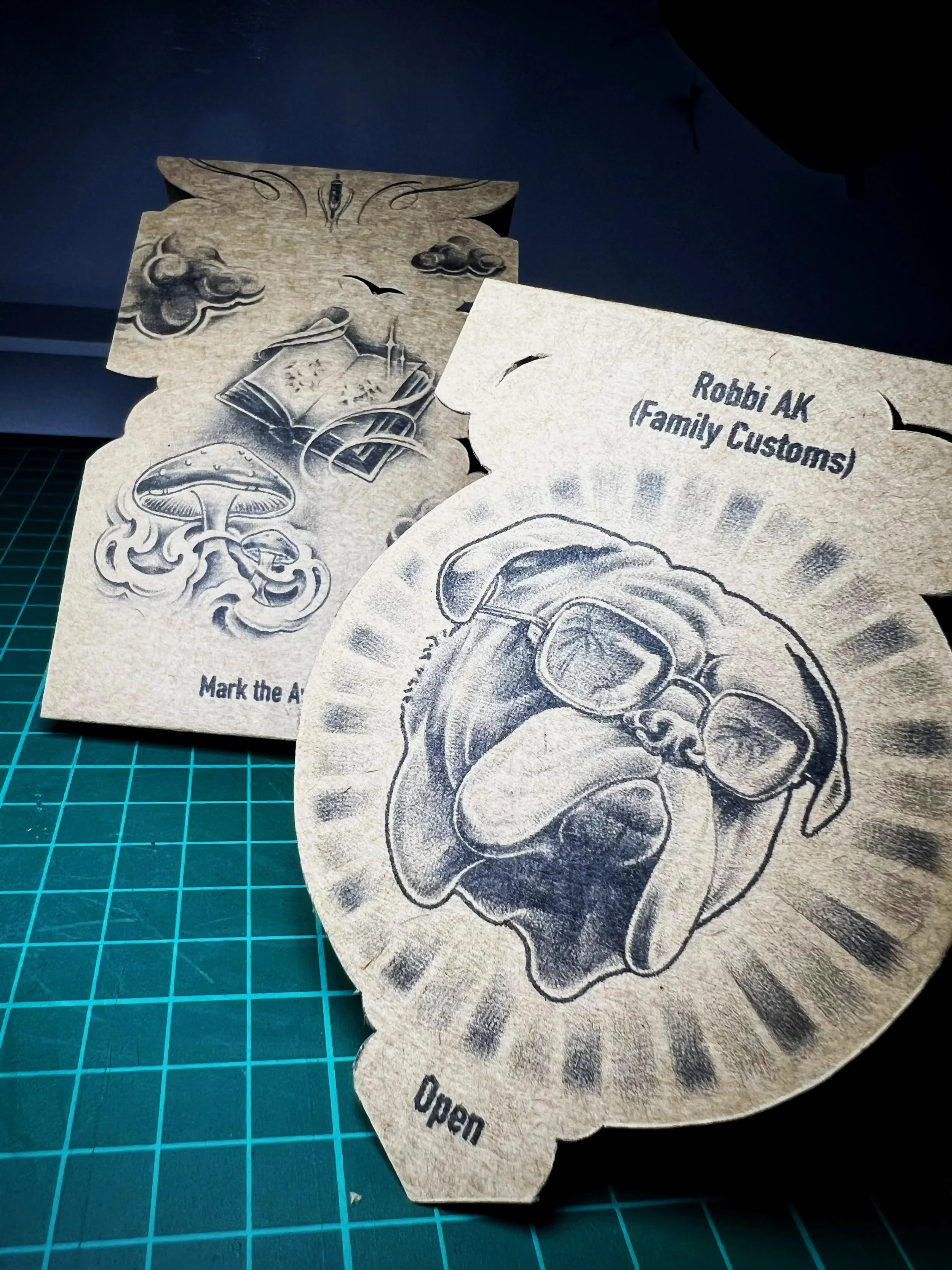 A set of paper bookmarks with engraved illustrations of a mushroom, a book, clouds, and bats on a green cutting mat.