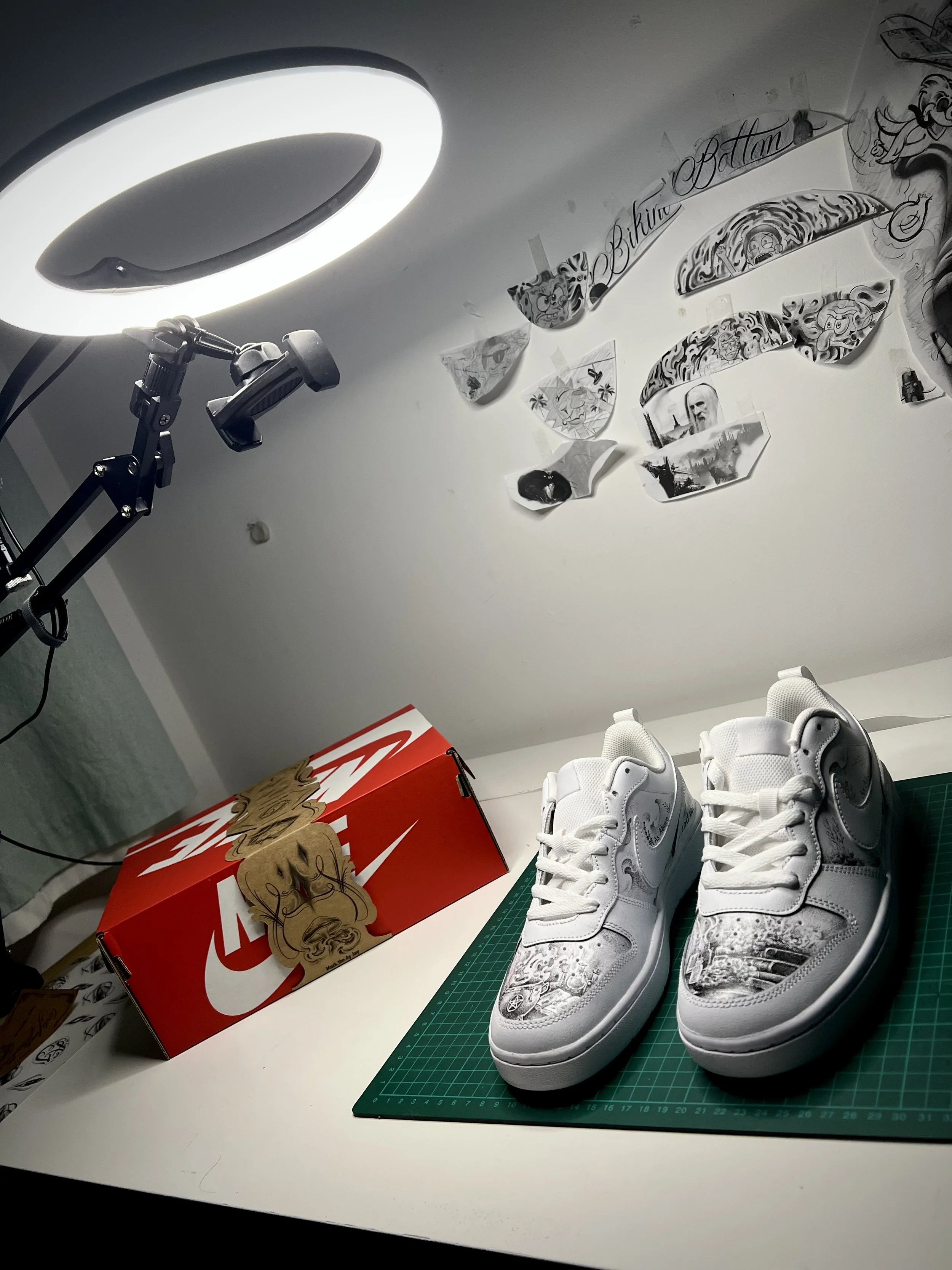 White sneakers with intricate designs placed on a green cutting mat on a white table, with a red Nike shoe box and paper tags nearby, under a large LED light.
