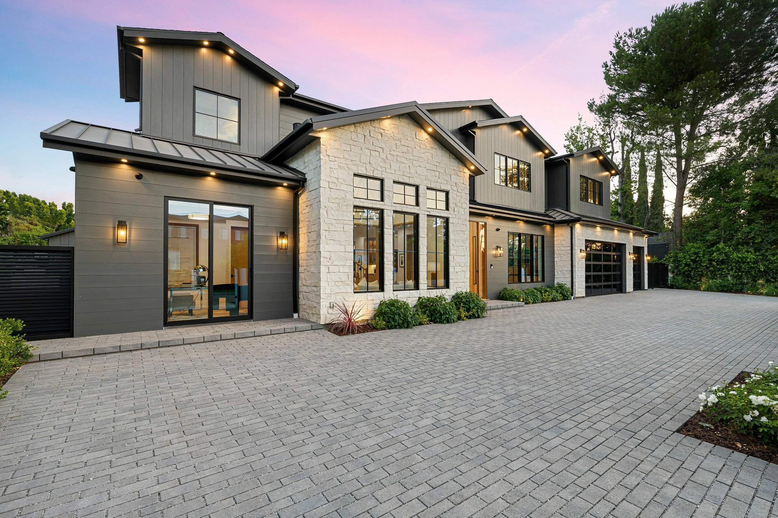 Modern house front with large windows, focus is on the windows, stone and siding exterior, landscaped small bushes, paved driveway, trees nearby, and sunset sky.