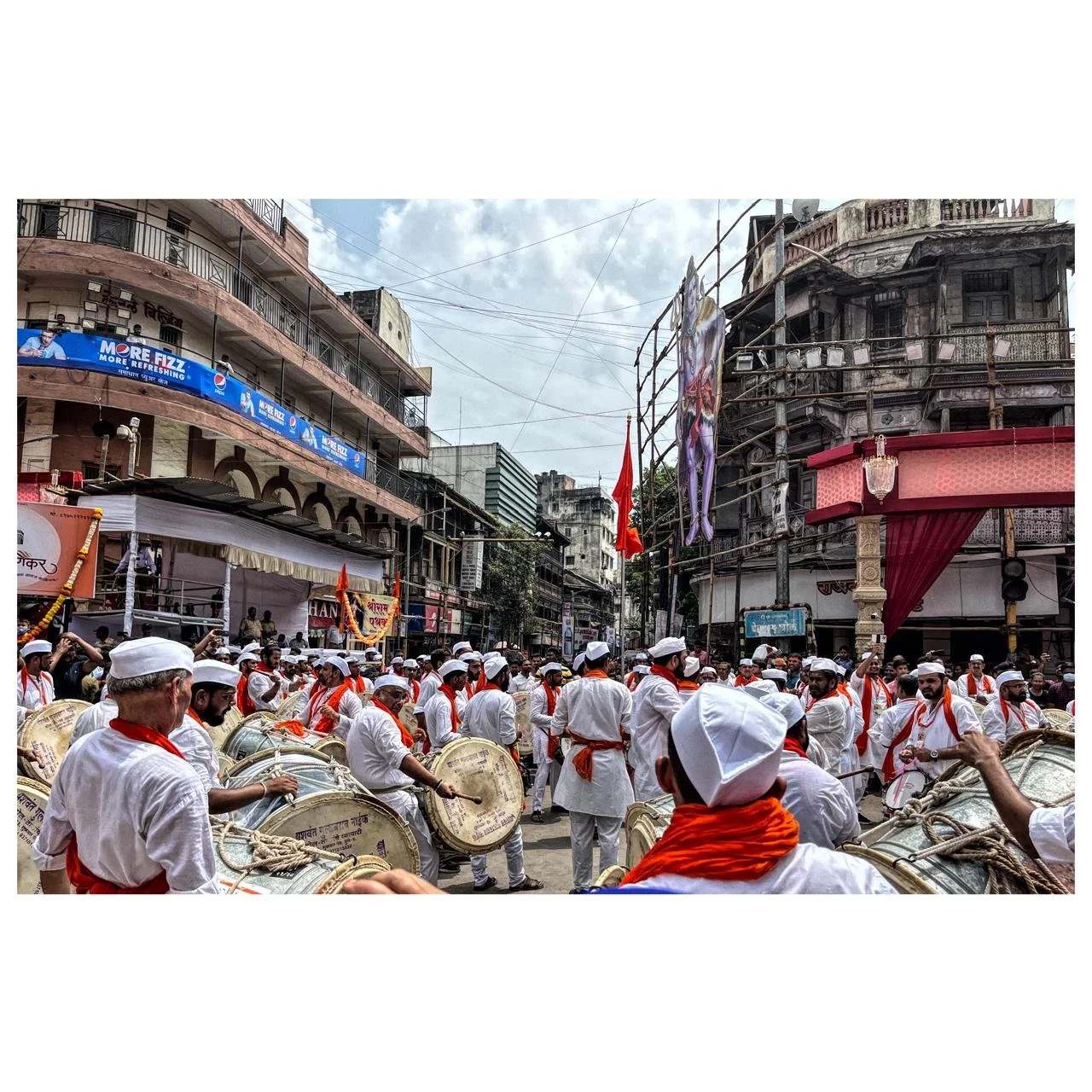 A street scene with a large group of people dressed in white with orange scarves and caps, participating in a parade or festival, with drums and musical instruments, in an urban area with old buildings, scaffolding, and banners visible.