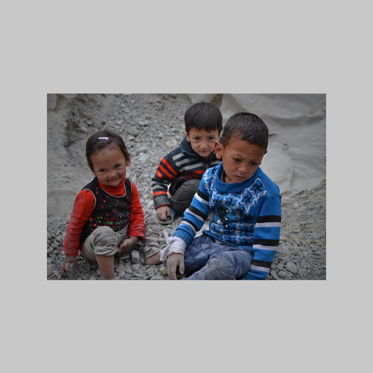 Three young children sitting on rocky ground in a rocky outdoor environment, wearing colorful sweaters and casual clothing.