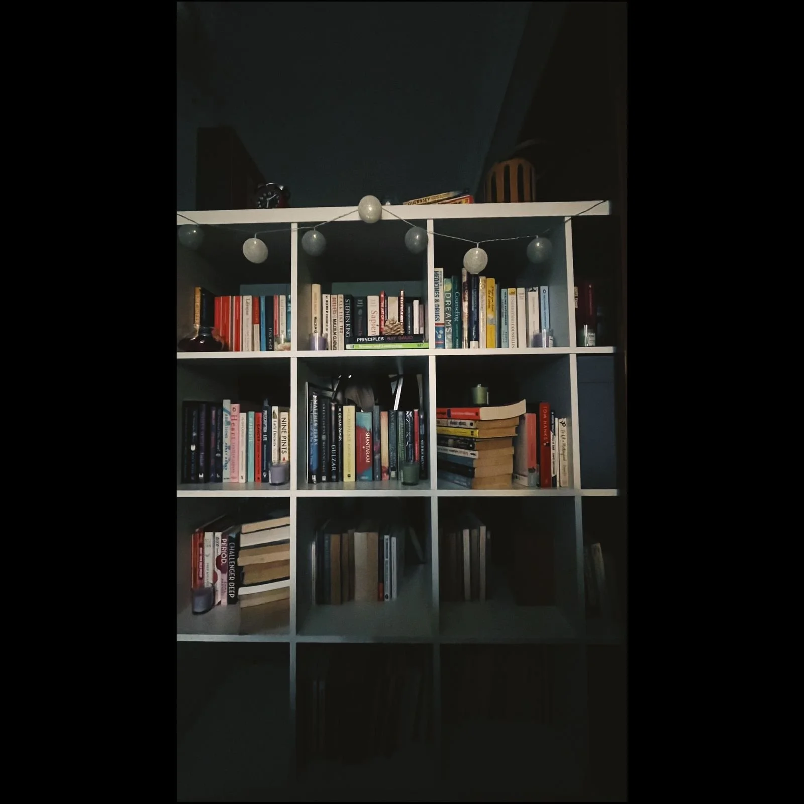 White bookshelf filled with books, decorated with string lights, on top are a clock, some books, and a wooden lantern. The room is dimly lit.