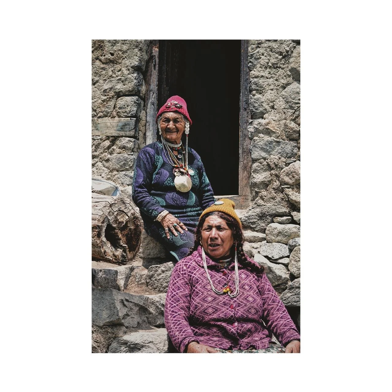 Two indigenous women sitting on stone steps in front of a stone building. The older woman wears a colorful sweater, beaded jewelry, a large pendant, and a pink hat, smiling. The younger woman wears a patterned pink shirt, yellow hat, braided hair, and colorful jewelry, with a serious expression.