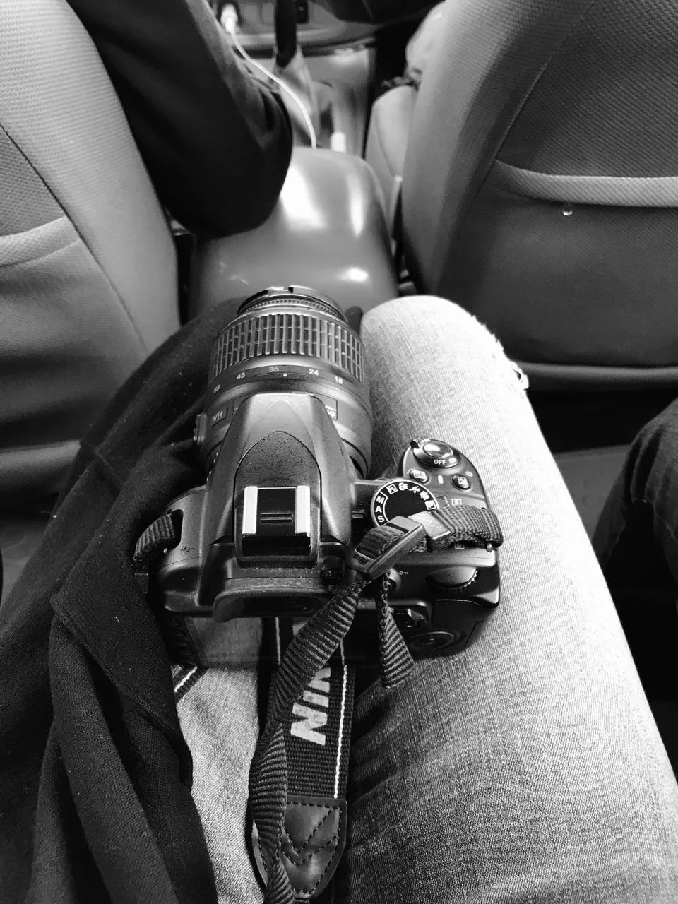 A DSLR camera resting on a person's lap inside a vehicle, with the view facing down the car's row of seats in black and white.