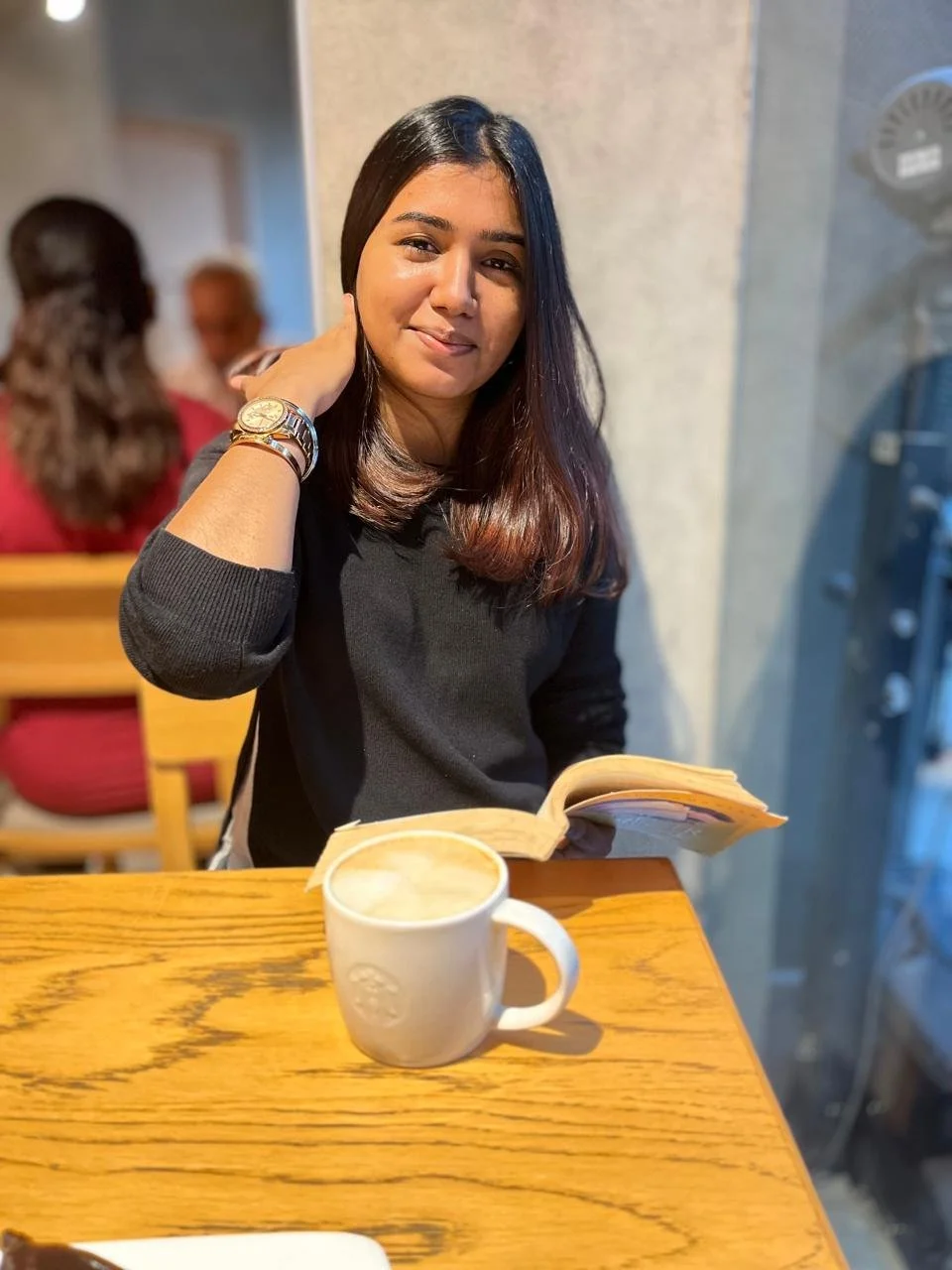 A young woman sitting at a wooden table in a coffee shop, holding an open book and smiling, with a cup of coffee in front of her.