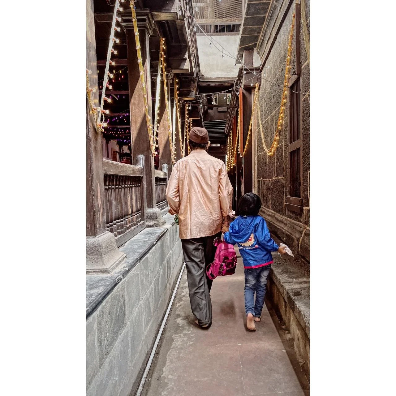 An adult and a child walk hand-in-hand down a narrow alley decorated with string lights and ribbons, with traditional dark stone buildings on either side.