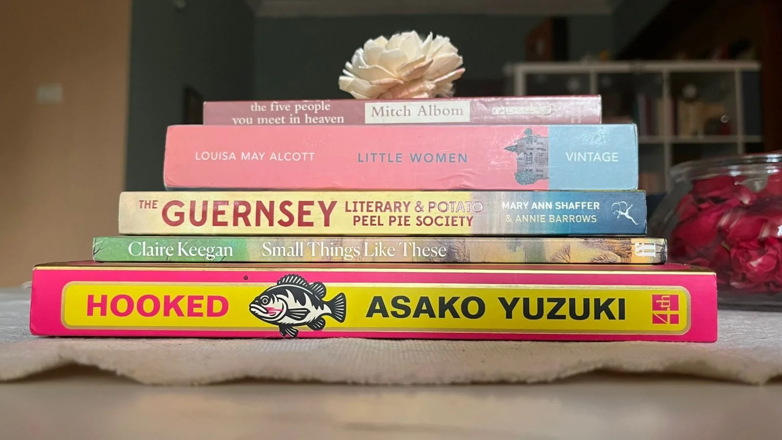 A stack of seven books arranged on a table, with a flower on top. The titles include 'The Five People You Meet in Heaven', 'Little Women', 'The Guernsey Literary and Potato Peel Pie Society', 'Small Things Like These', 'Hoked', and the top book by Mitch Albom.