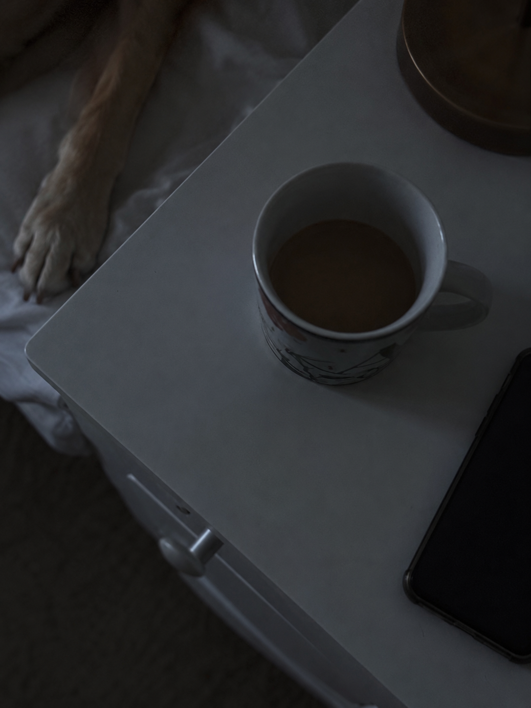 Top view of a coffee mug filled with coffee on a white nightstand, with a dog lying nearby and partially visible in the background.
