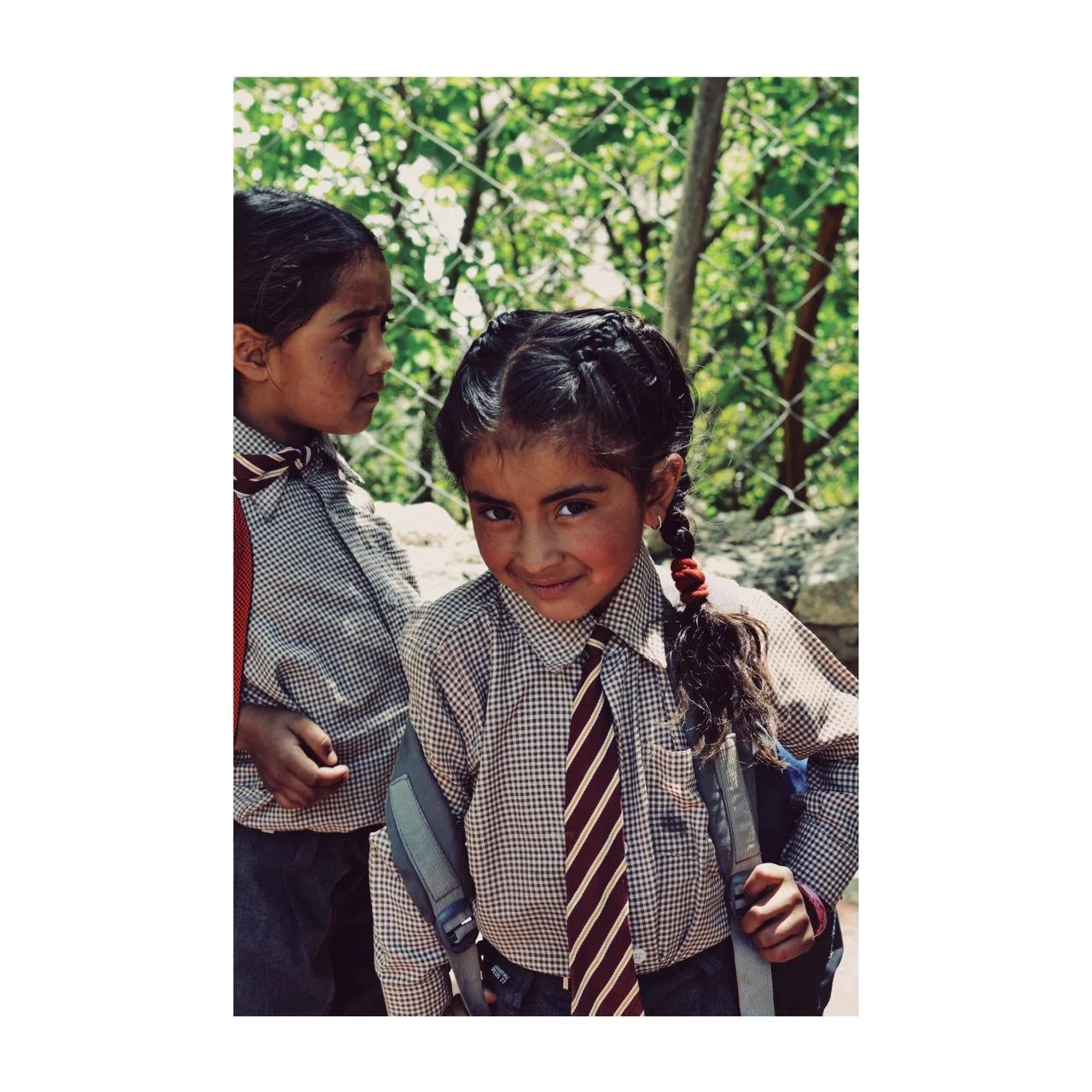 Two young girls wearing school uniforms with checkered shirts, striped ties, and backpacks, standing outdoors near a chain-link fence with green foliage in the background.