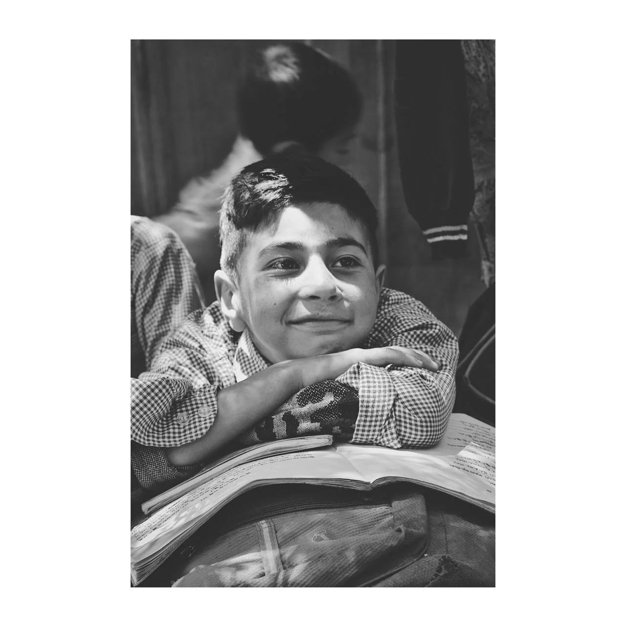 A young boy with short hair rests his head on his arms, smiling, with an open book in front of him. The photo is in black and white.