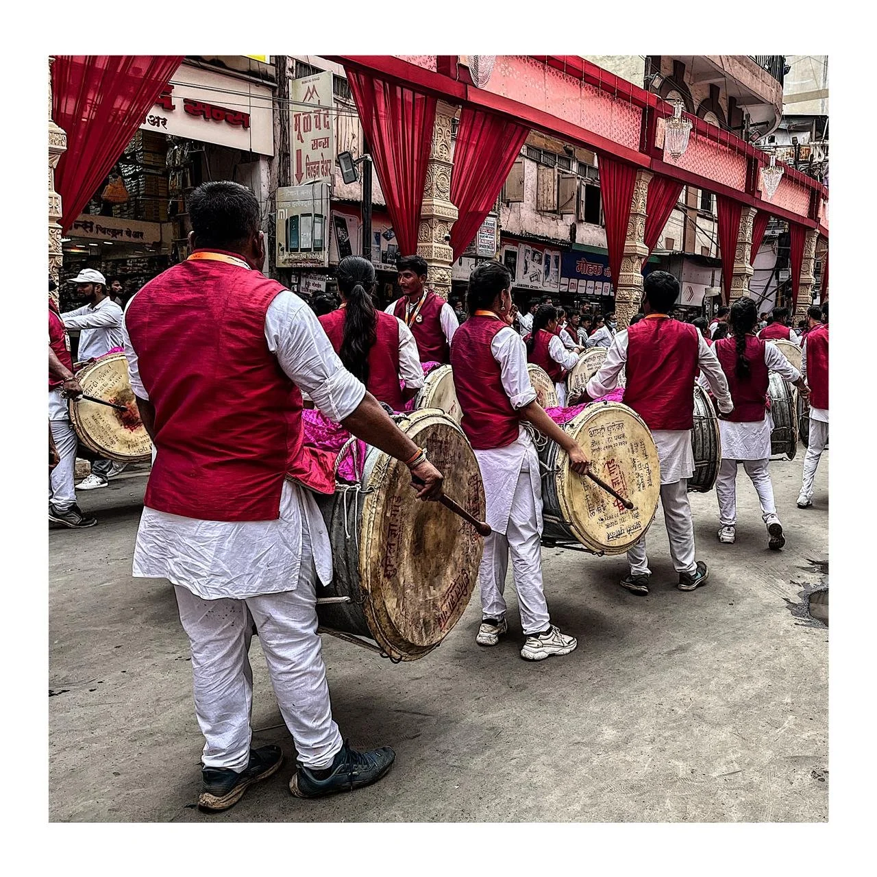 Group of traditional drummers in white and red outfits performing in a street parade with red drapes and a crowded urban background.