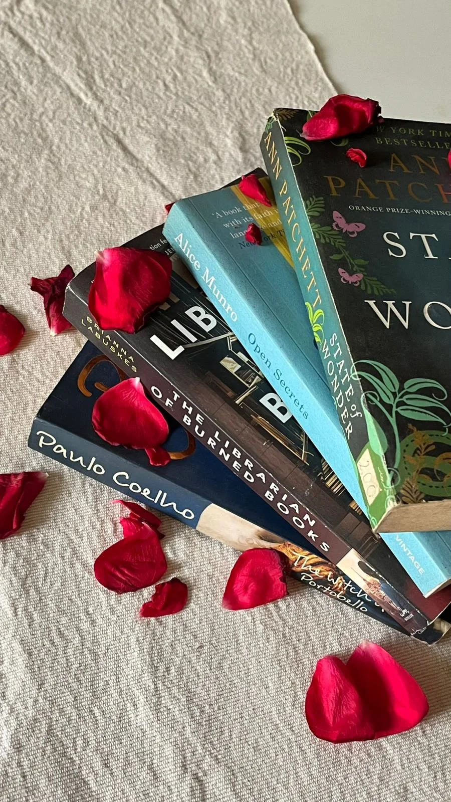 Stack of books on a beige textured surface with red rose petals scattered around.