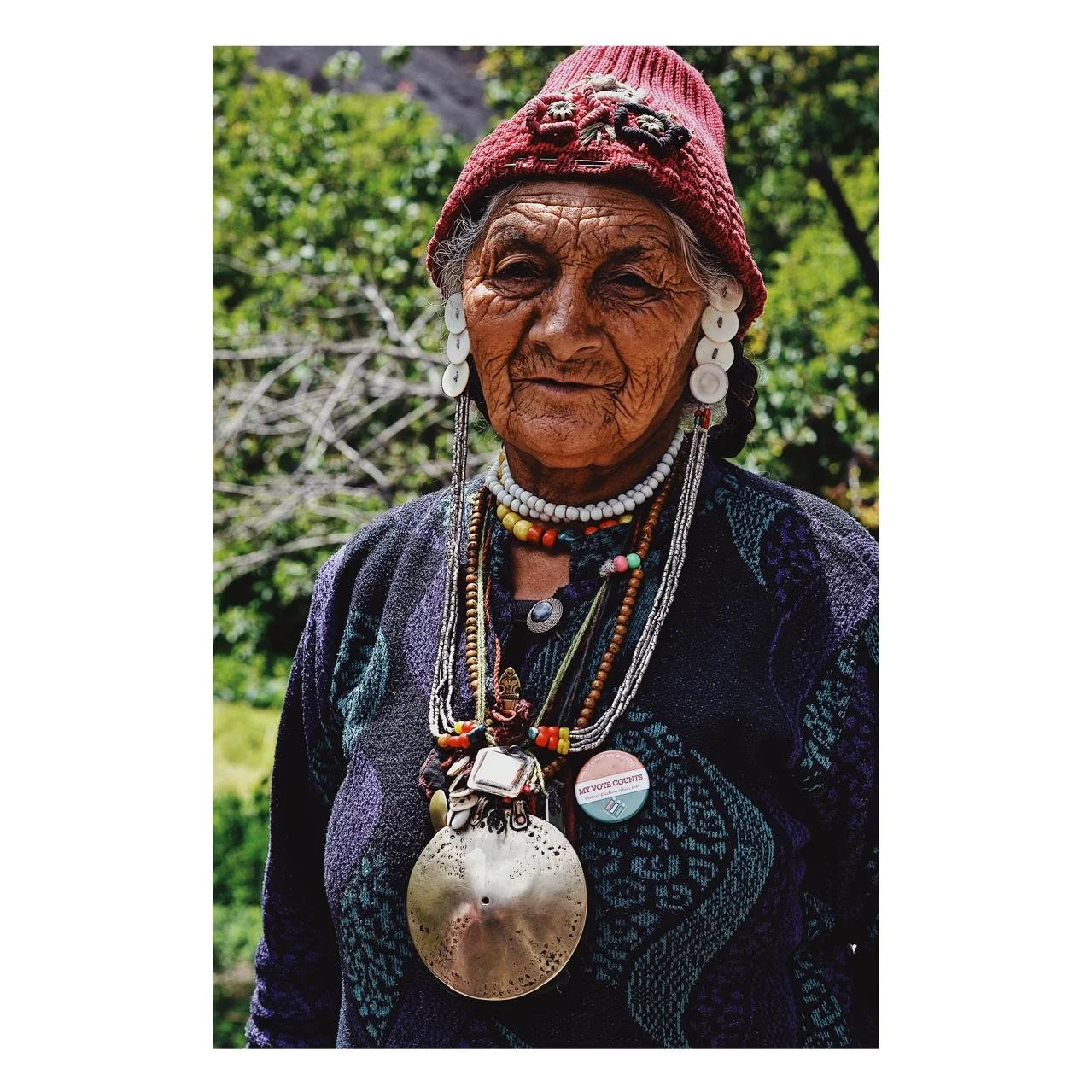 An elderly woman with dark skin and deep facial wrinkles, wearing a red knit hat with decorative pins, large earrings, and multiple layered beaded necklaces. She is dressed in a dark, patterned garment and stands outdoors with green foliage in the background.