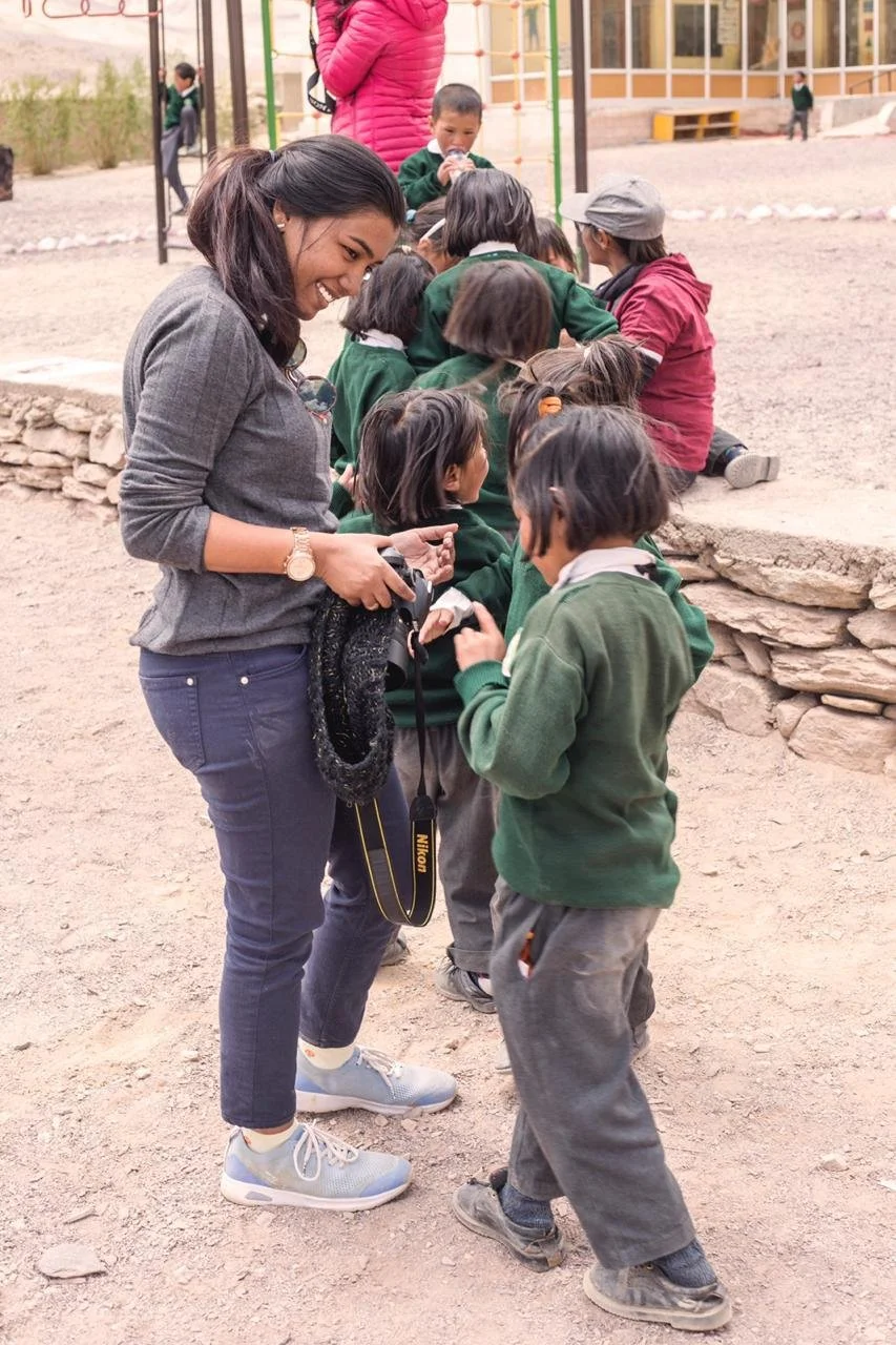 A woman and several children in a line at an outdoor playground. The woman is smiling and holding a camera, engaging with a child in front of her. The children are wearing green and gray uniforms, and some are wearing hats. In the background, there is a climbing structure and other children.