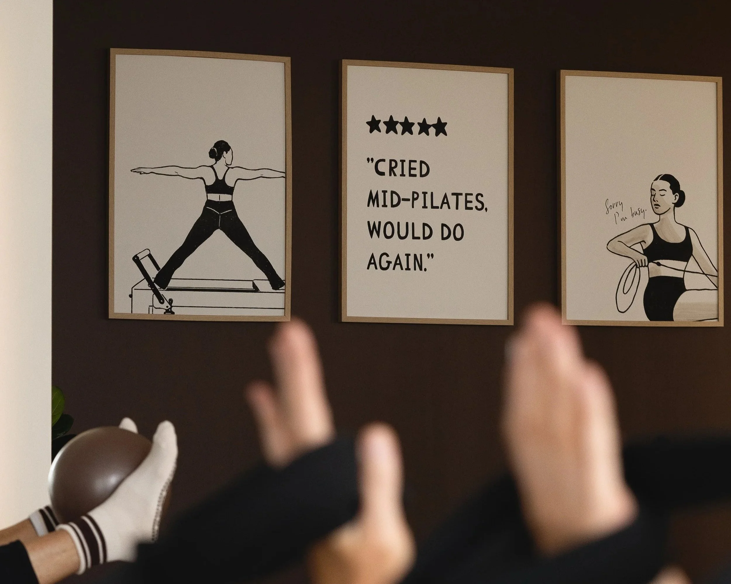 Framed fitness-themed wall art featuring Pilates illustrations and a five-star quote, viewed above a seated person