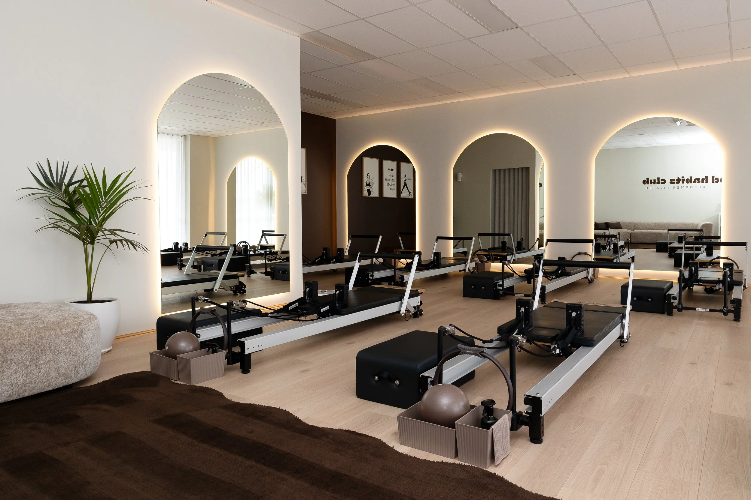 Bright Pilates studio with multiple reformer machines, arched backlit mirrors, wood flooring, and a potted palm