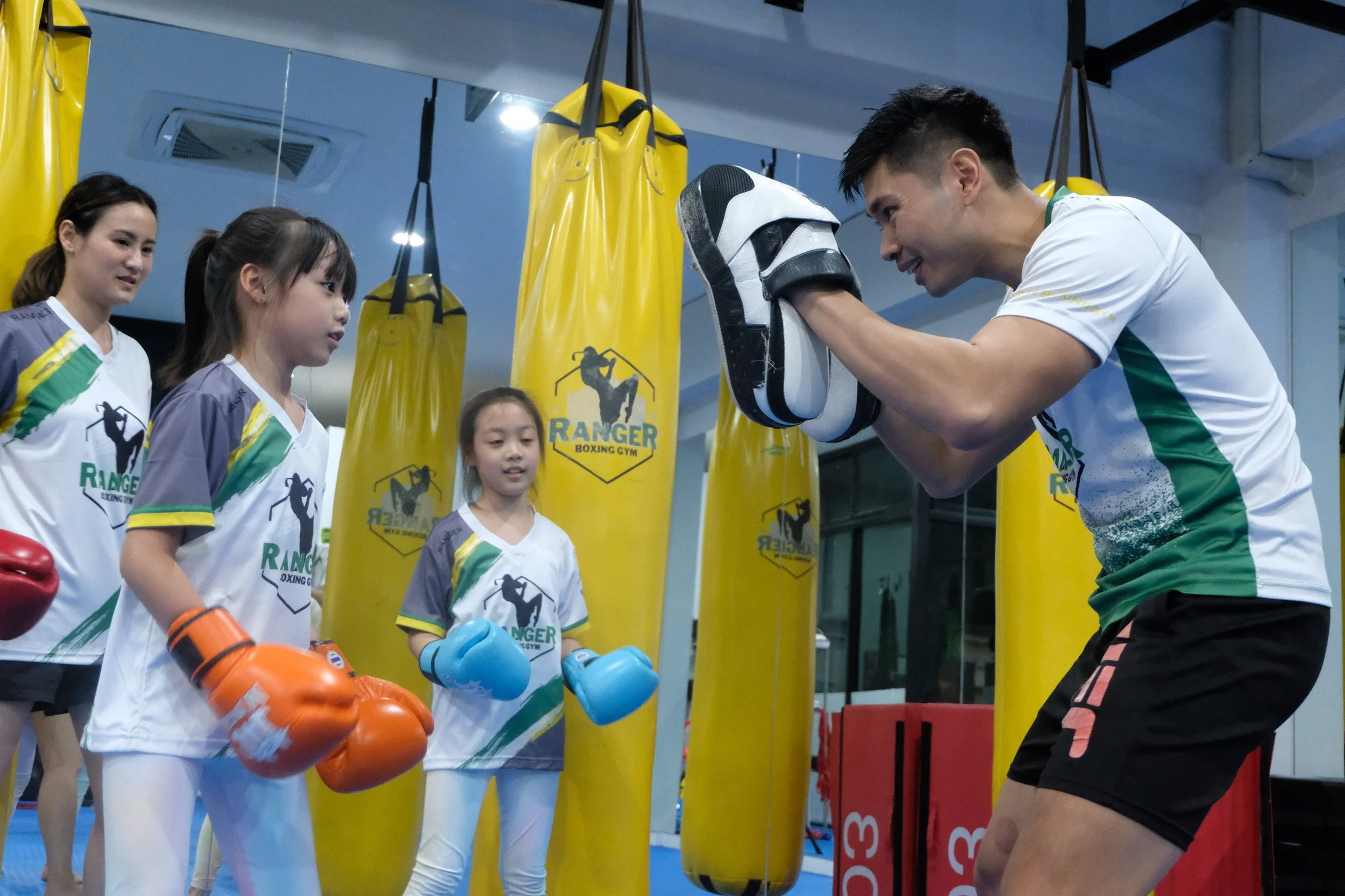 A group of young female boxers in boxing gear listening to a male trainer in a gym, with yellow punching bags and boxing equipment around them.