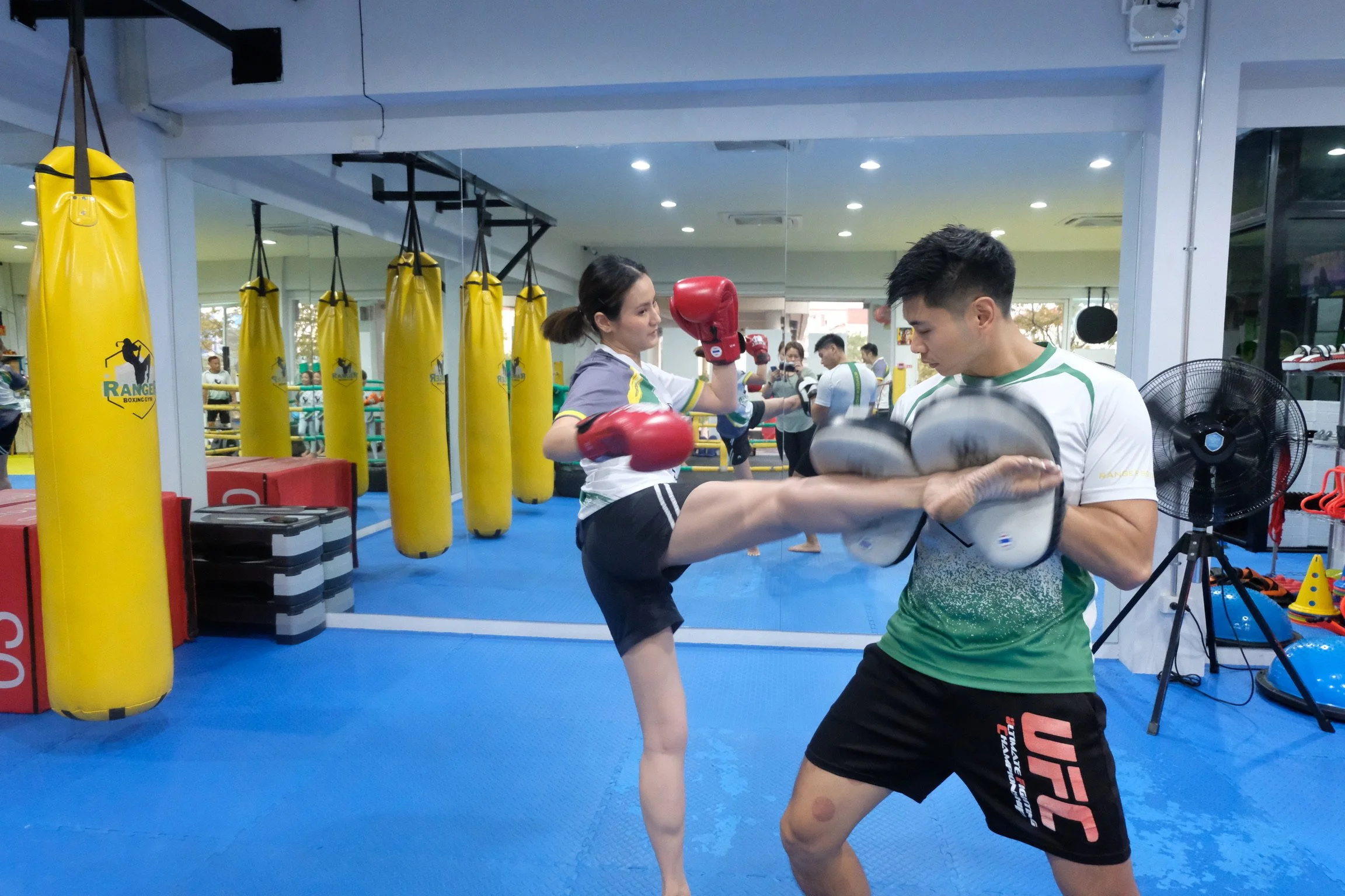 A woman practicing kickboxing with a trainer in a boxing gym. The woman is wearing red boxing gloves and kicking while the trainer assists with her form. The gym has yellow punching bags and a blue mat.