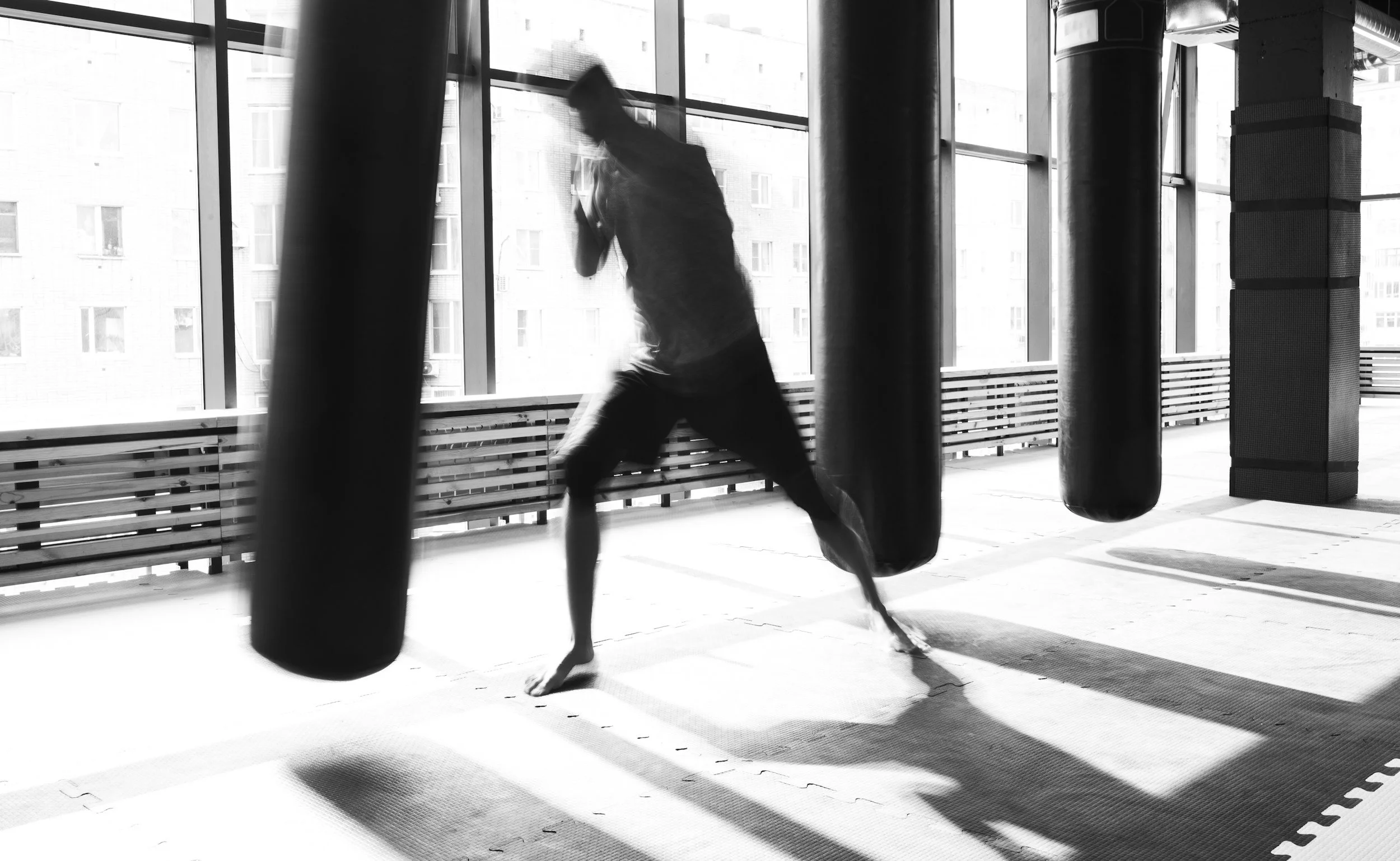 A person in a gym practicing martial arts or boxing, seen through a window with punching bags hanging in the foreground.