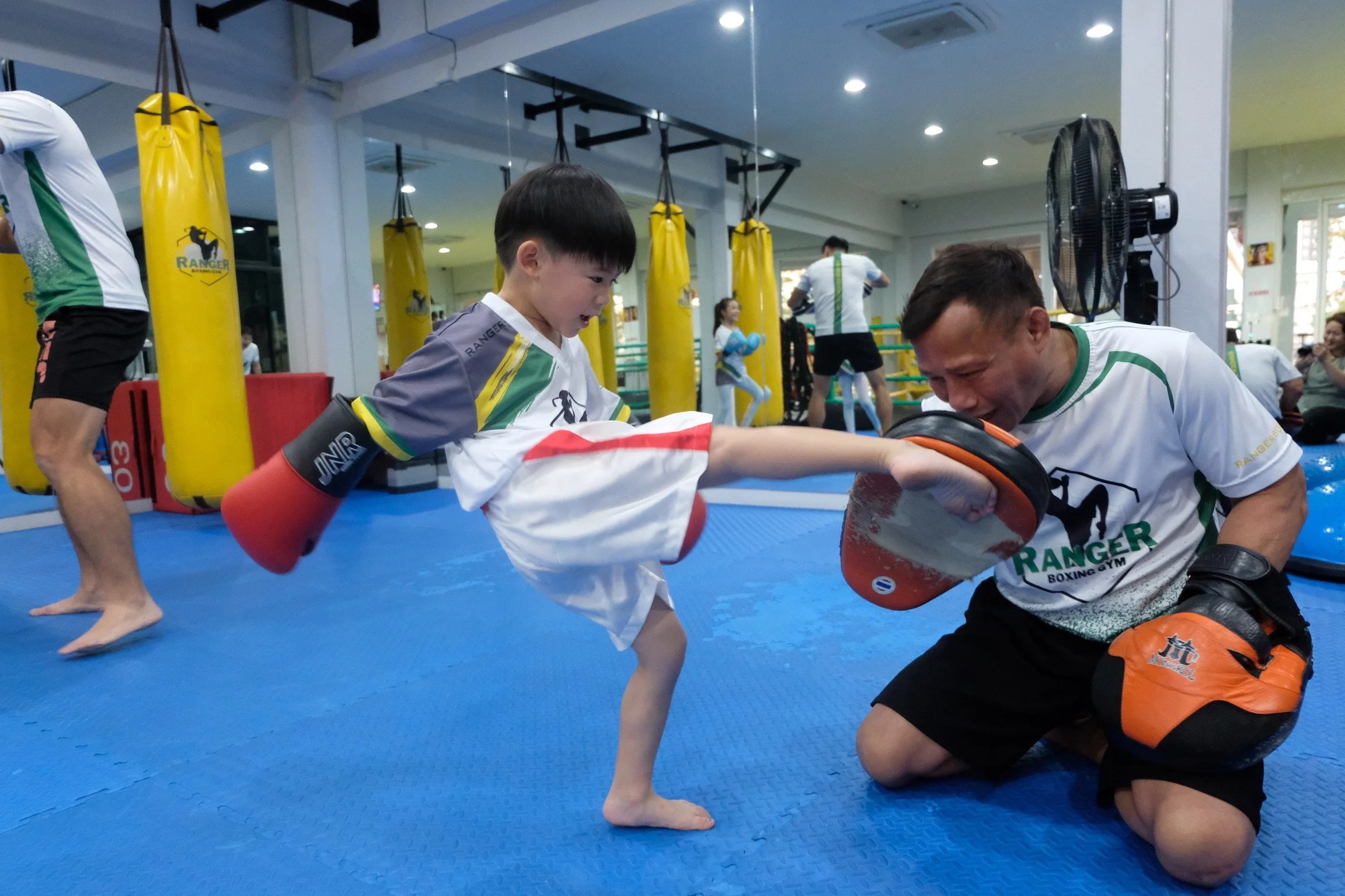 A young boy practicing boxing on a punching bag with a coach or trainer in a gym. The gym has yellow punching bags and several people in the background.